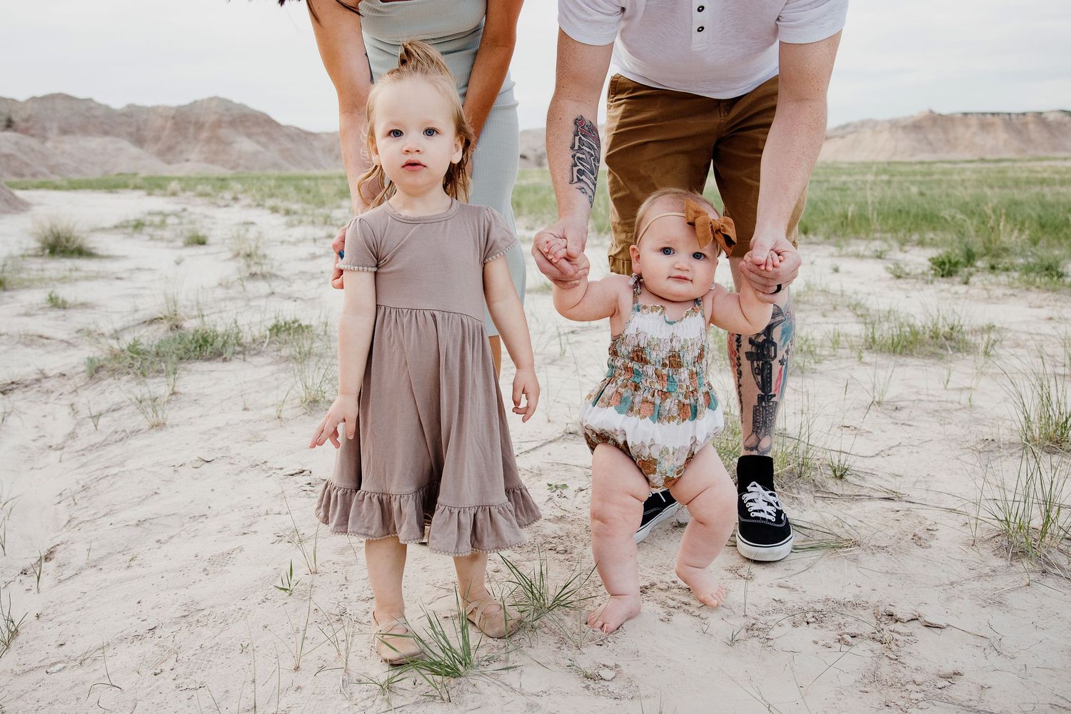 Two young sisters holding parents hands standing in sandy desert landscape with badlands backdrop.