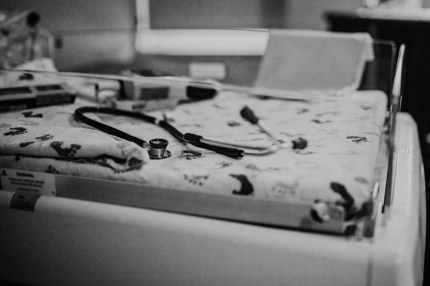 Black and white photo of a stethoscope resting on a baby crib with patterned bedding in a hospital nursery.