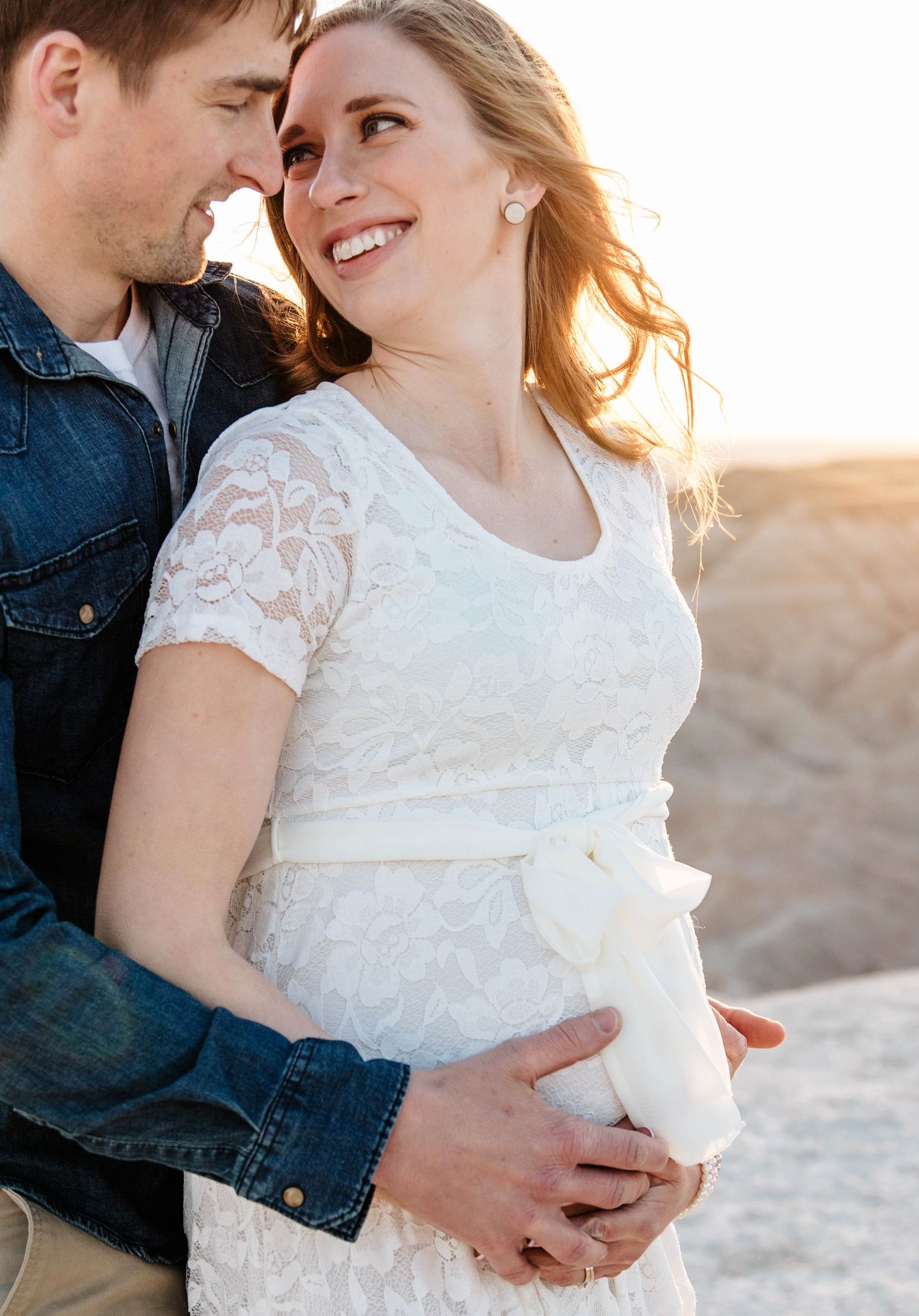 Smiling pregnant woman in white lace dress embraced by partner at golden hour in Badlands.