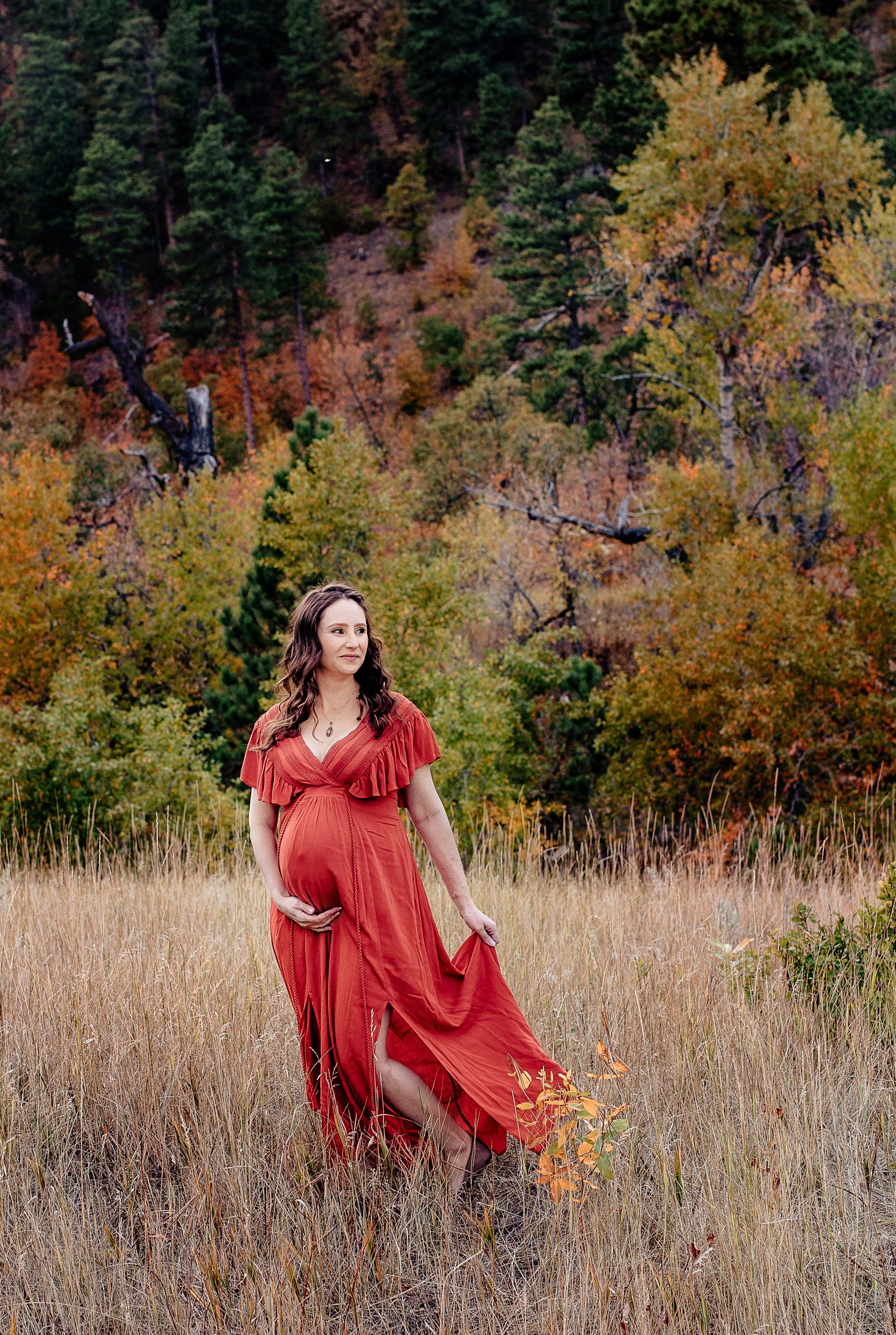 Pregnant woman in red flowing maternity gown walks through autumn field holding wildflowers.
