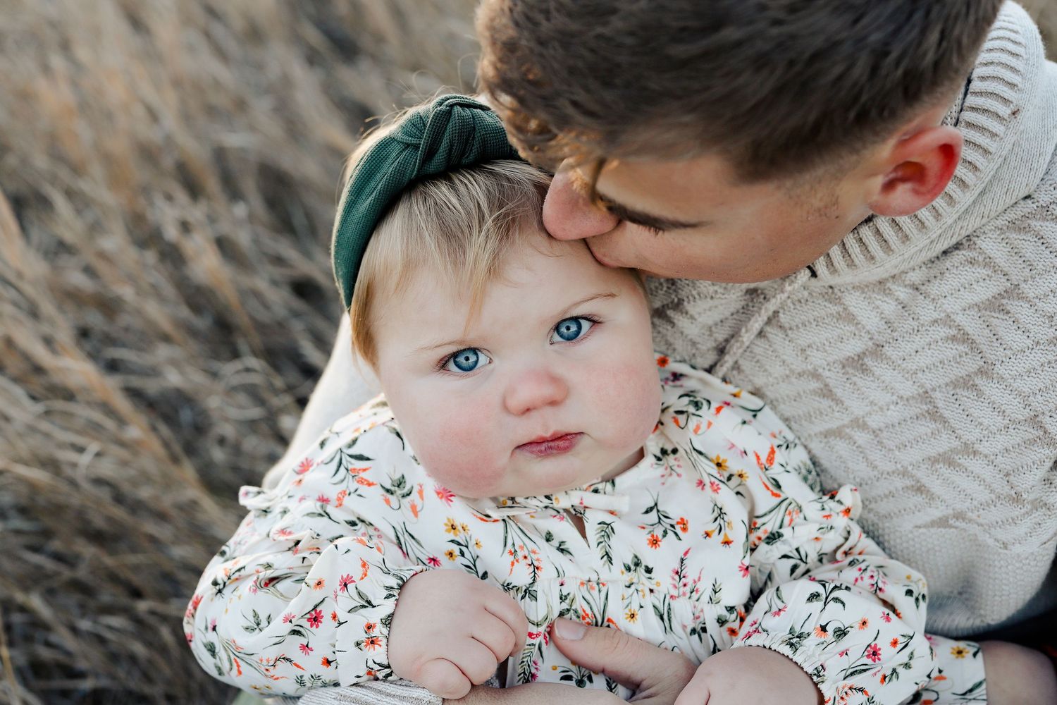 Father gently kissing his blue-eyed baby girl on the cheek in a warm outdoor portrait.