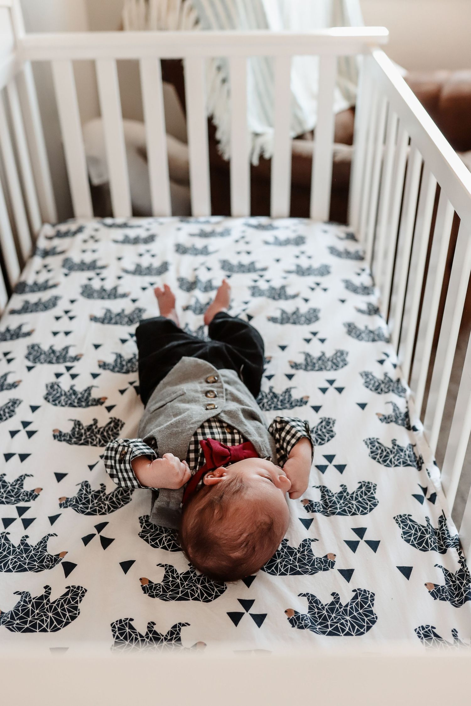 Newborn baby in gray outfit lying on back in white crib with black and white crown patterned sheet.