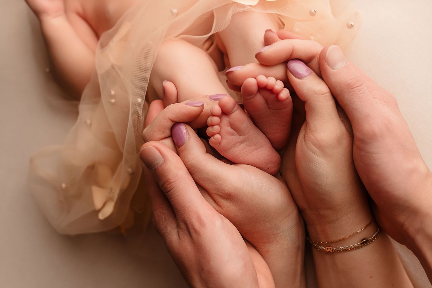 Two parents gently cupping tiny newborn baby feet together in soft peach fabric background.