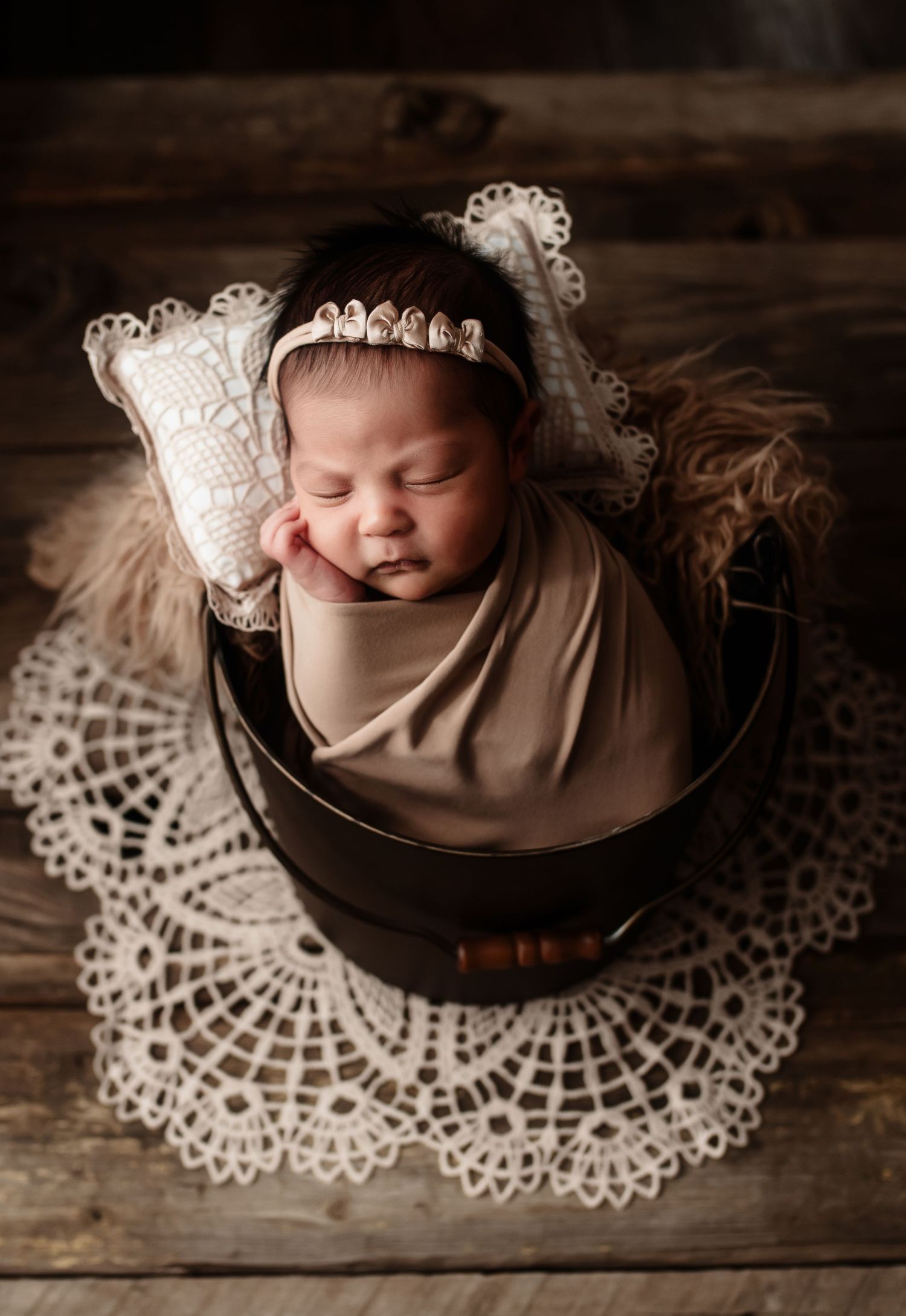 Sleeping newborn baby swaddled in beige wrap sitting in wooden bucket on lace doily.