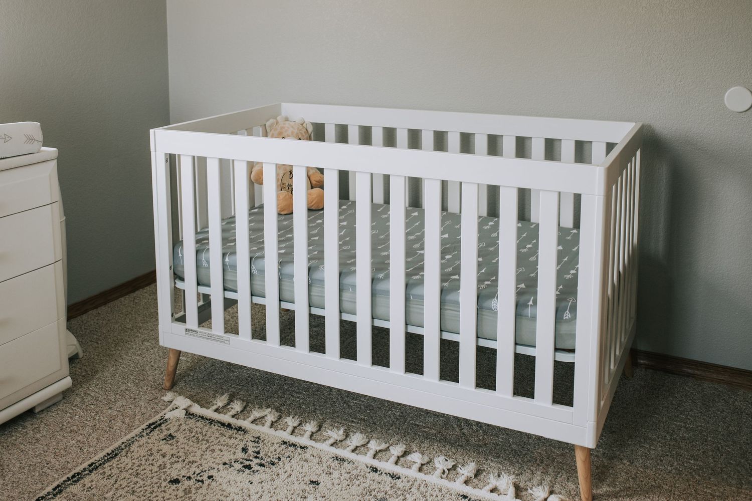 Modern white wooden baby crib with arrow-print sheet in a soft gray nursery room.