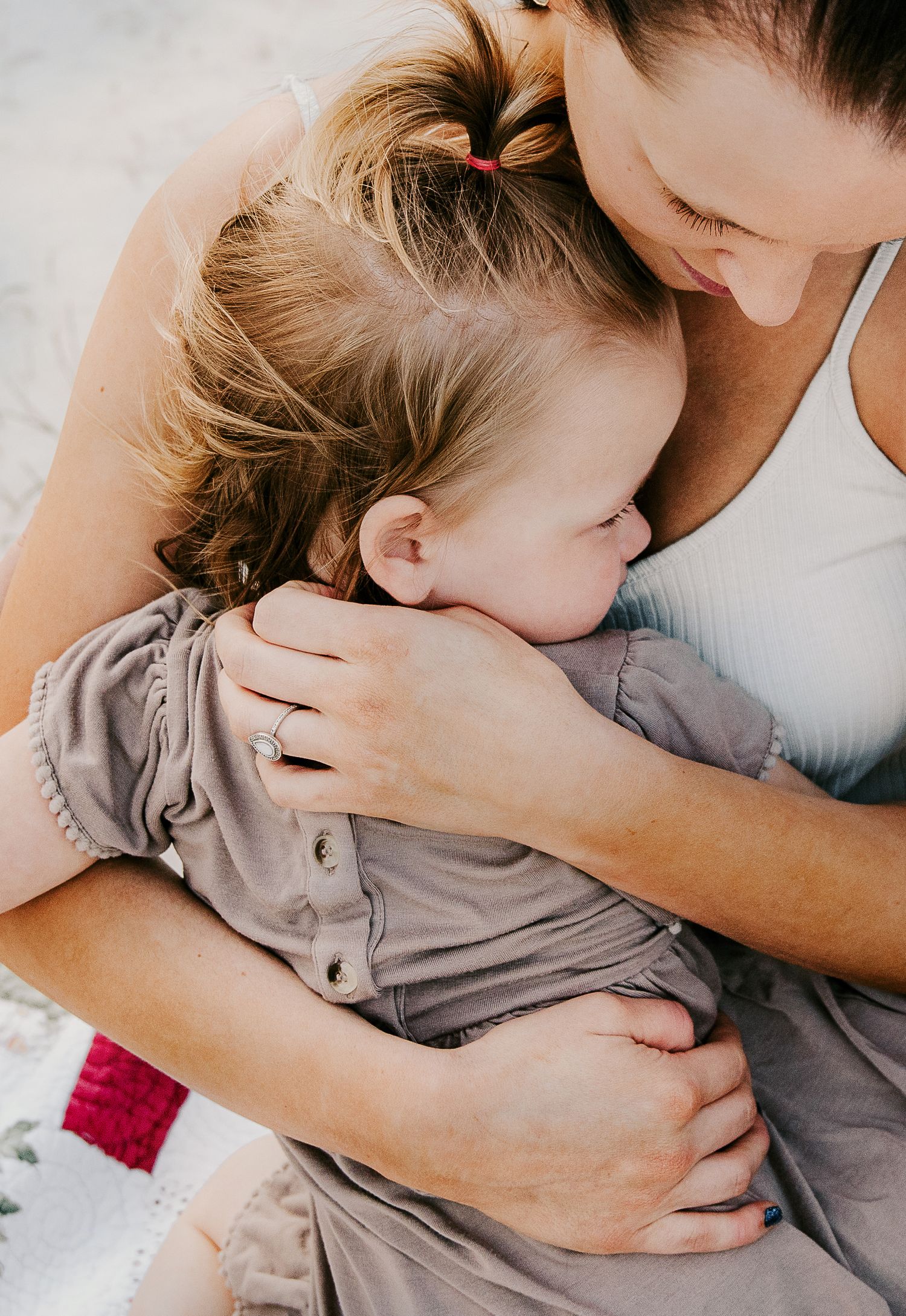 Mother tenderly hugging young daughter wearing gray dress on a sandy beach setting.
