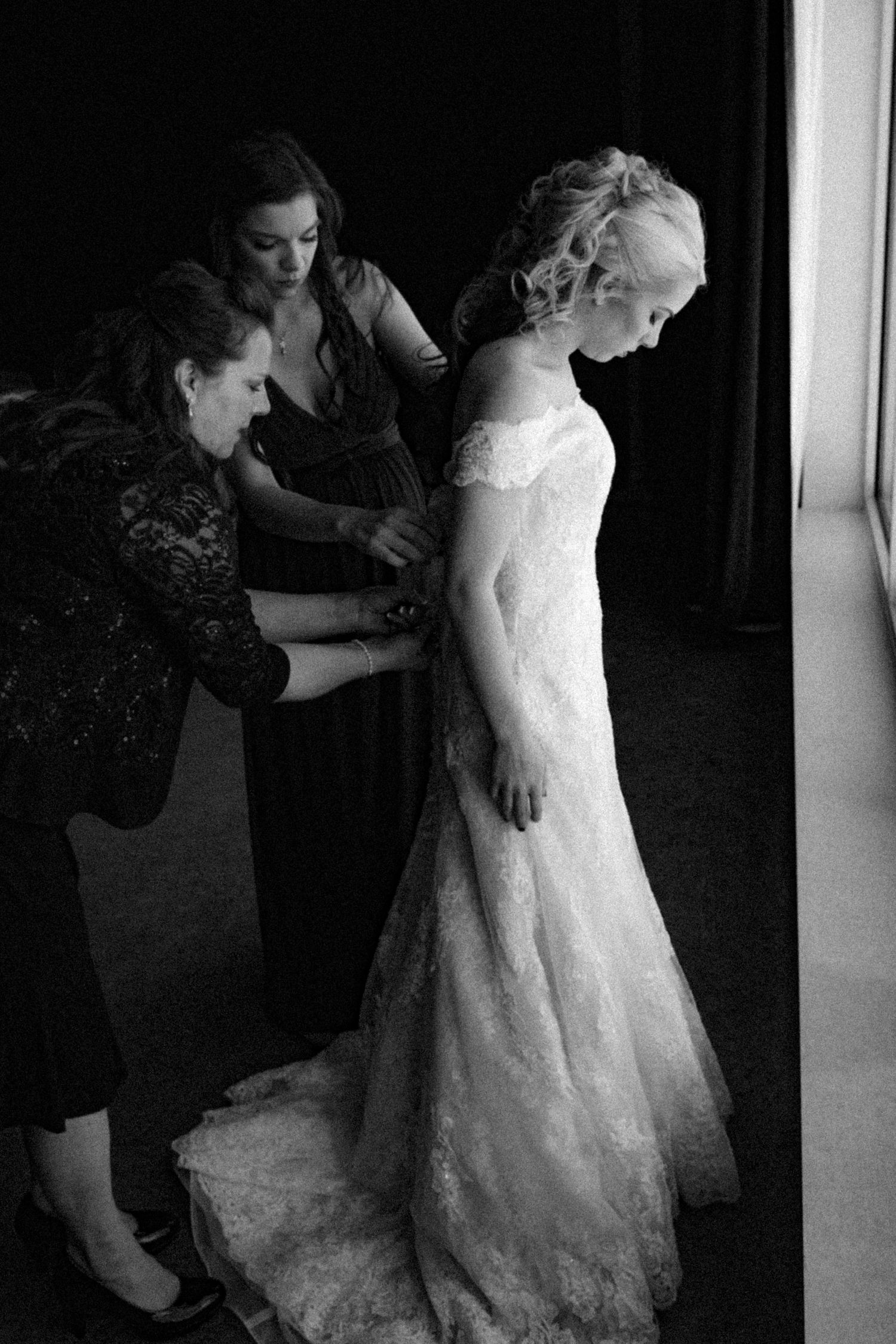 Two bridesmaids help a bride in a lace wedding gown get dressed in a black and white wedding photo.