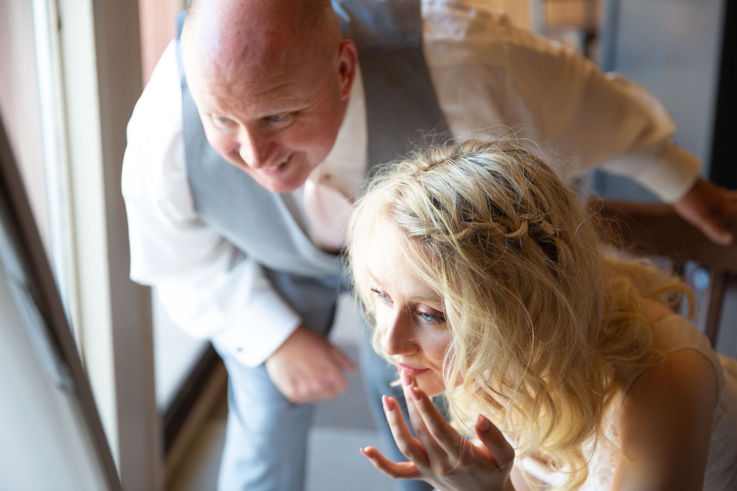 Bald man in gray suit leaning over blonde woman in white dress, both looking out a window at a wedding.