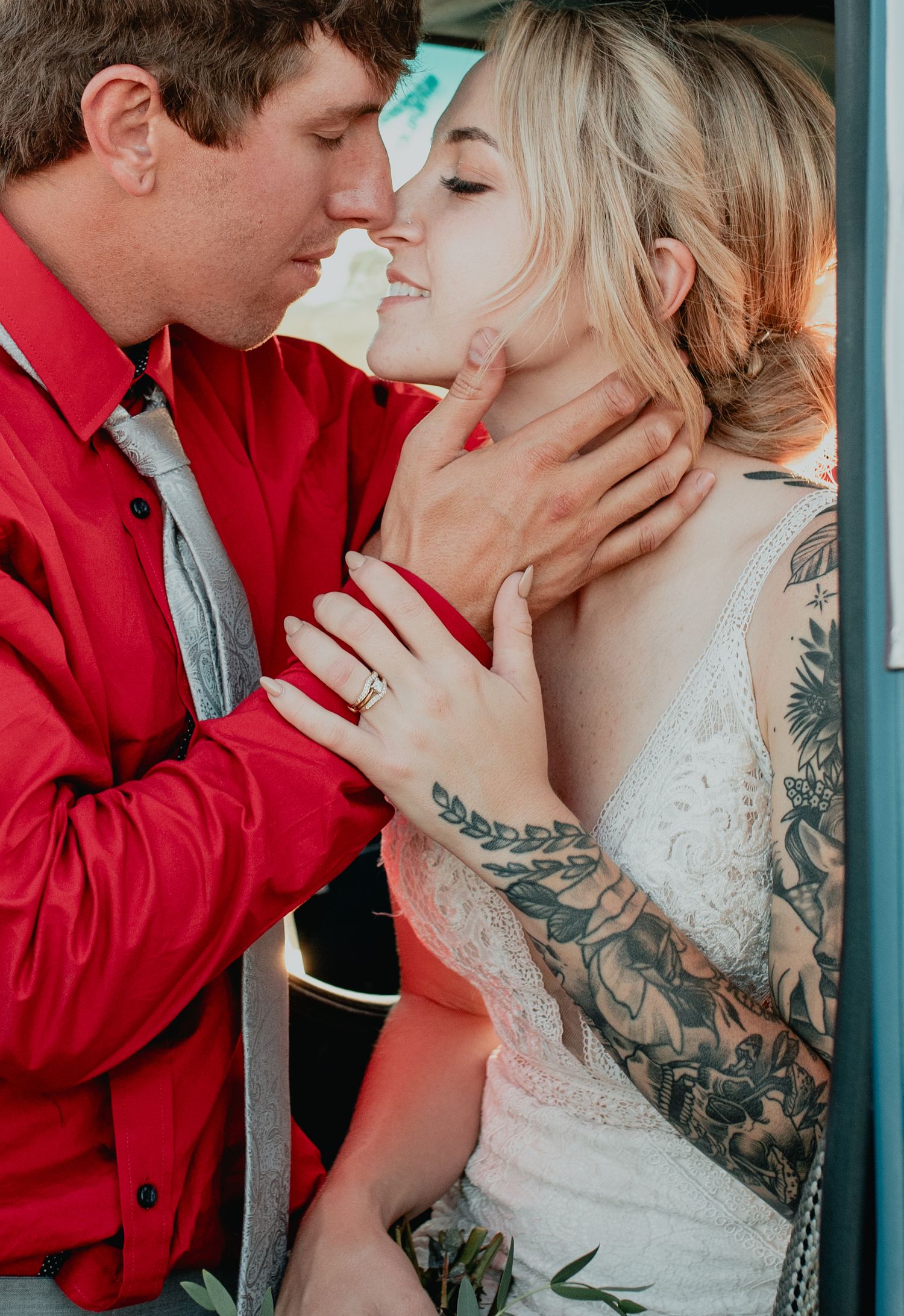 Bride with floral tattoo sleeve and groom in red shirt sharing an intimate moment, wedding rings visible.