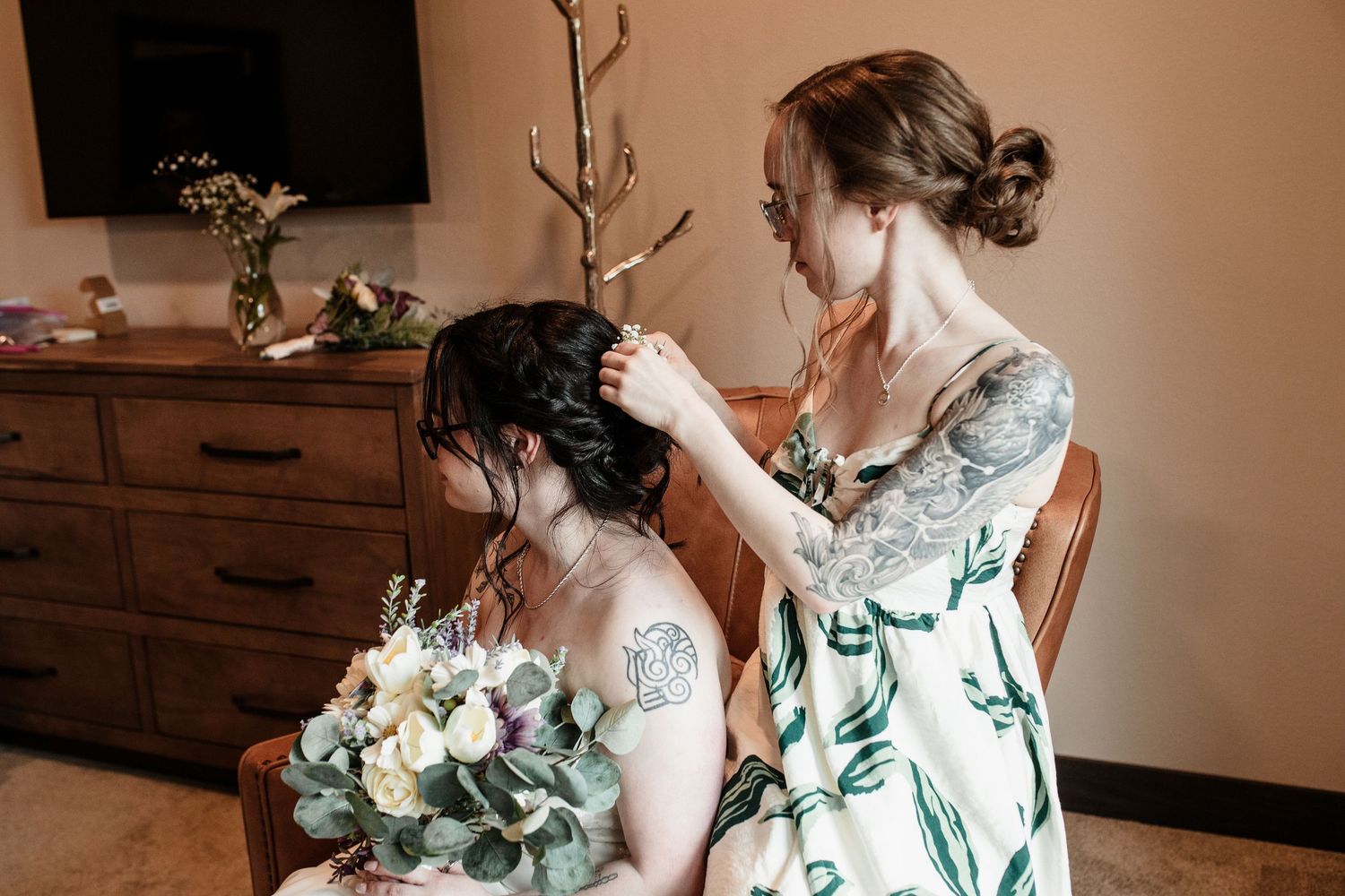 Bridesmaid with tattoos adjusting bride's updo hairstyle while bride holds a white and green wedding bouquet.