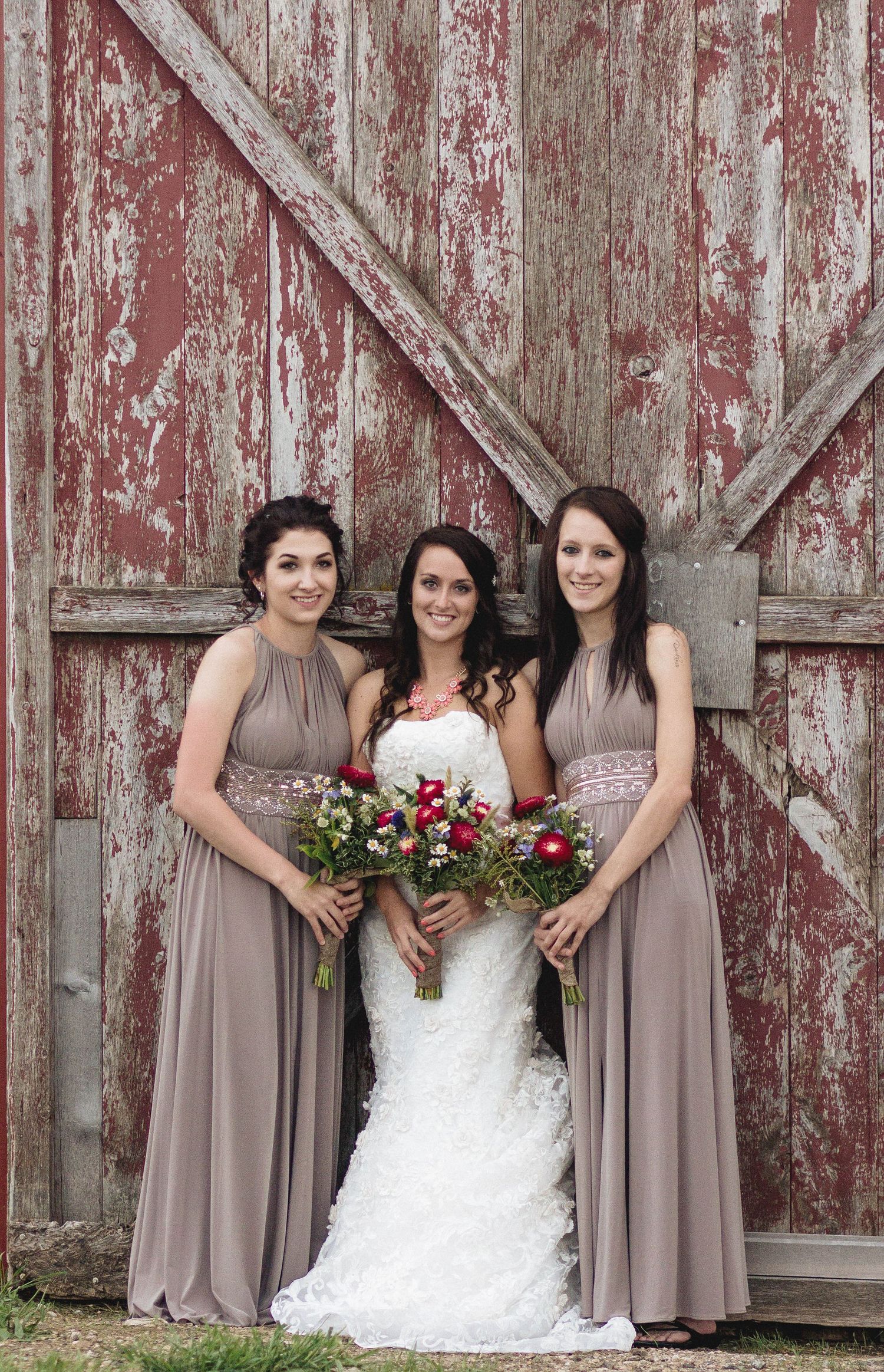 Bride in white lace gown flanked by two bridesmaids in taupe dresses holding red bouquets before rustic barn doors.