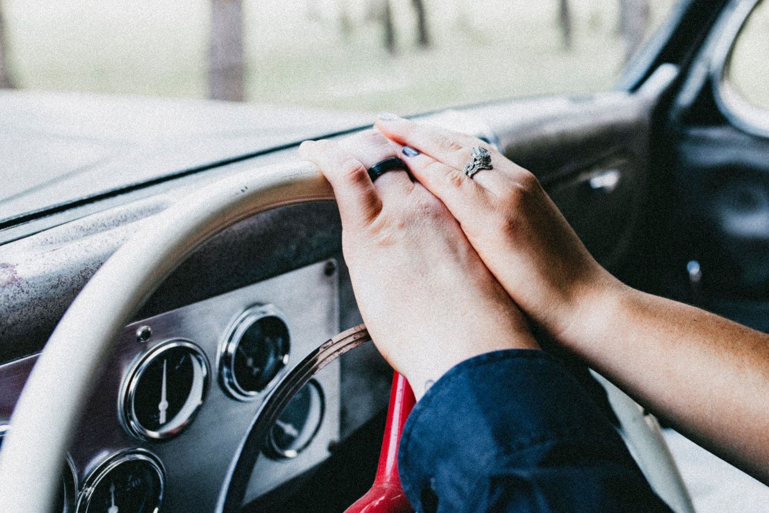 Two hands with rings clasped together over a vintage car steering wheel and dashboard.