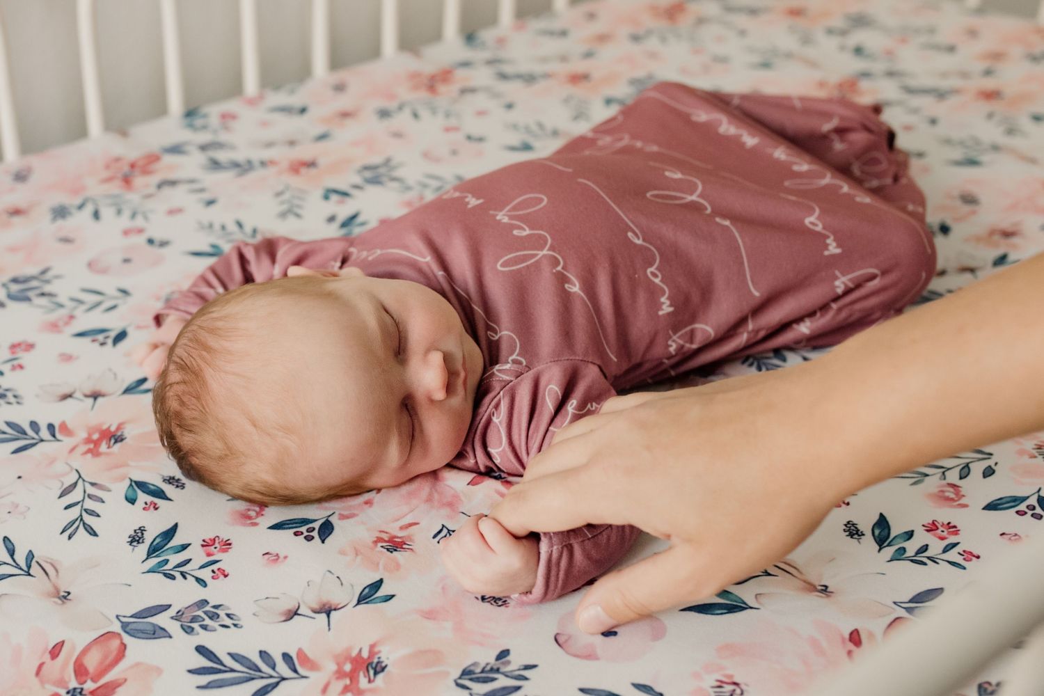 Newborn baby in dusty rose swaddle lying in floral crib with parent's hand gently holding them.