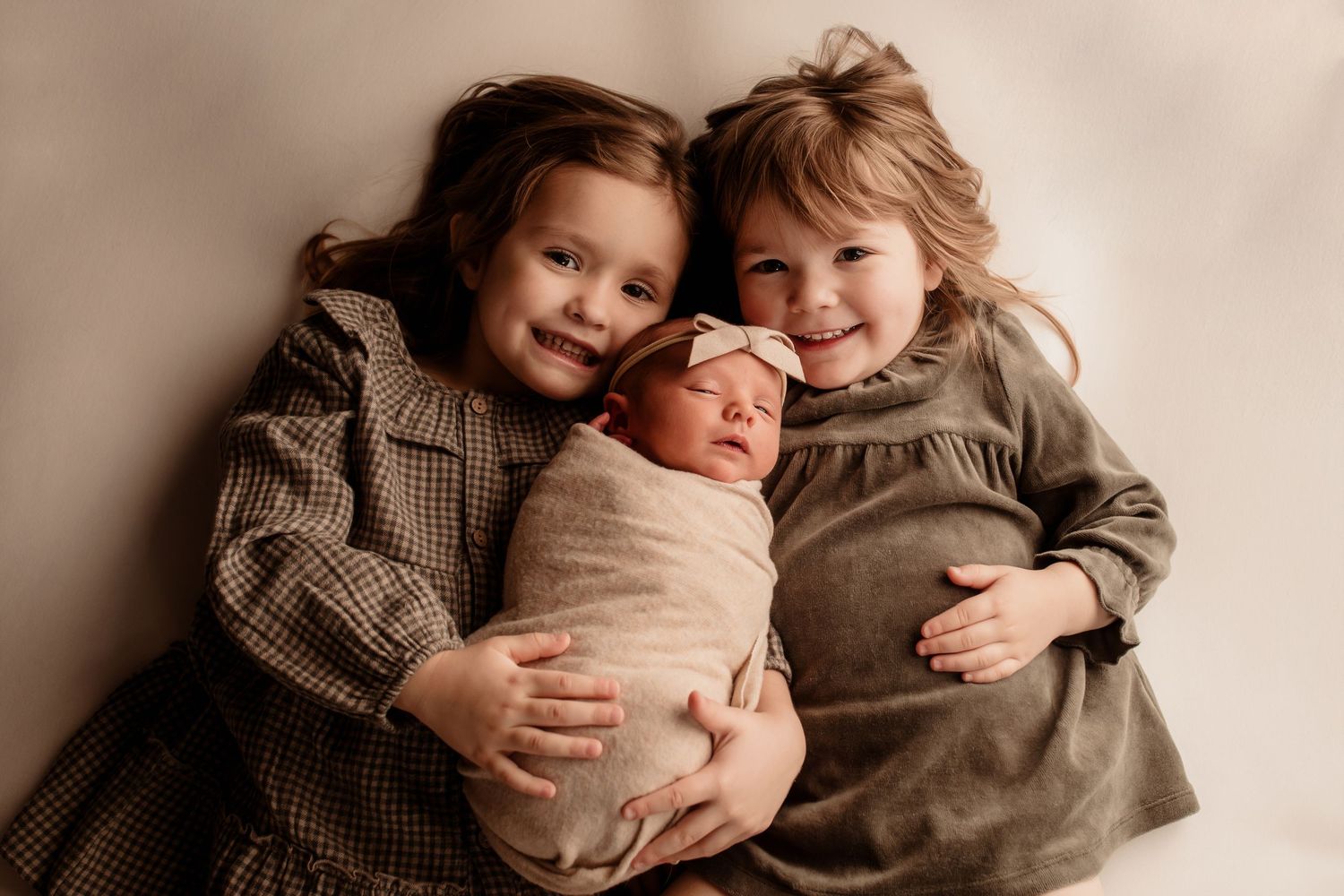 Two young sisters in dresses smiling while holding swaddled newborn baby together.