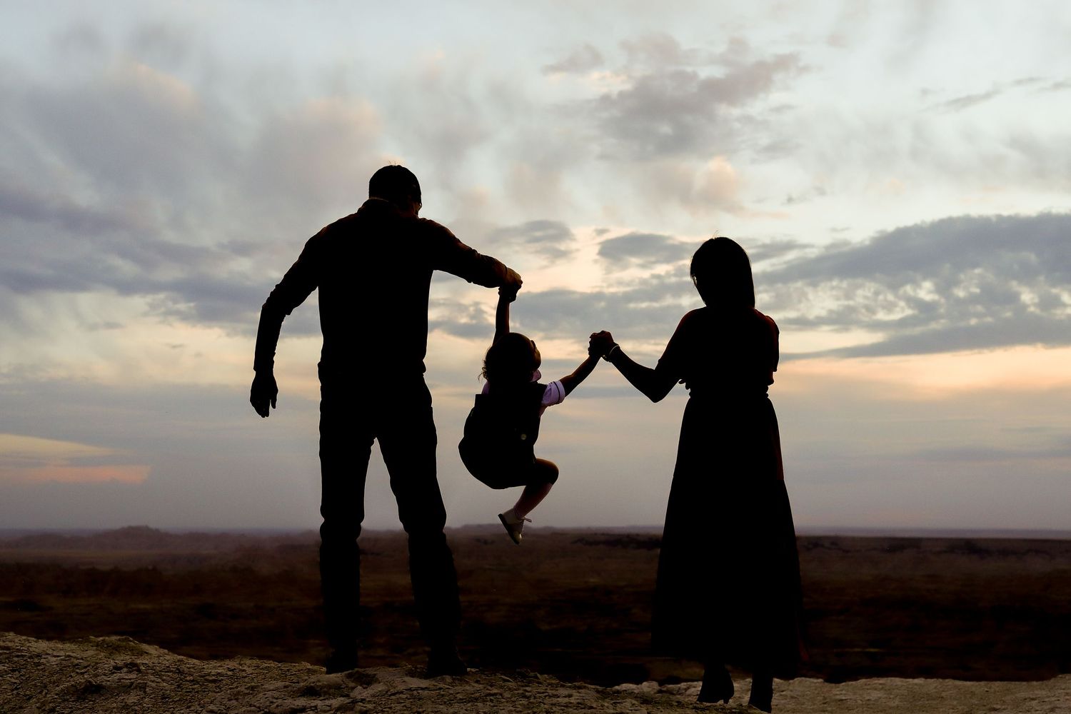 Silhouette of family swinging child between parents at sunset with dramatic cloudy sky backdrop.