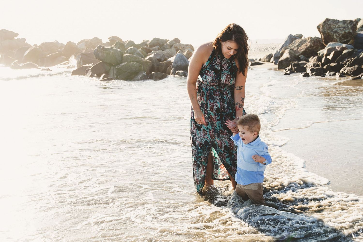 Mother in floral dress helping young toddler boy wade through shallow ocean waves at beach.