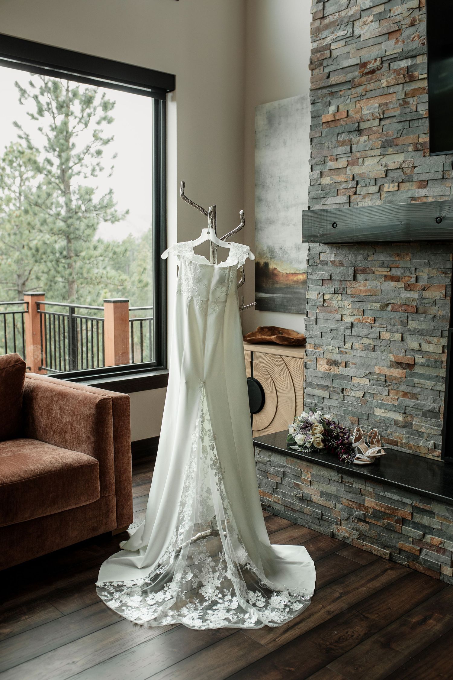 Elegant white wedding dress with lace hem hanging on antler hooks near stone fireplace and large window in rustic venue.