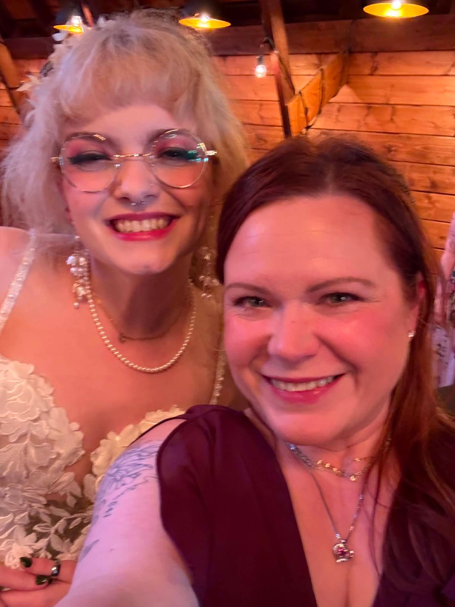 Two smiling women pose together at a wedding celebration, one in a white lace bridal gown with pearls, the other in burgundy.