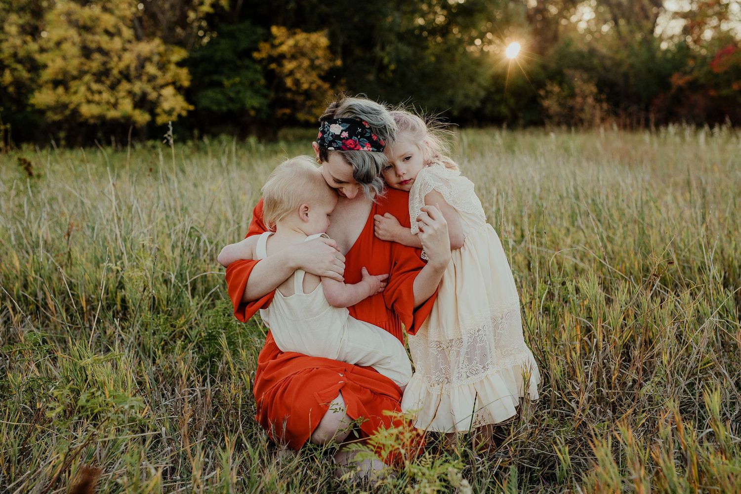 A mother in a red dress sits in a golden field at sunset, tenderly holding her baby while a young girl in white leans in.