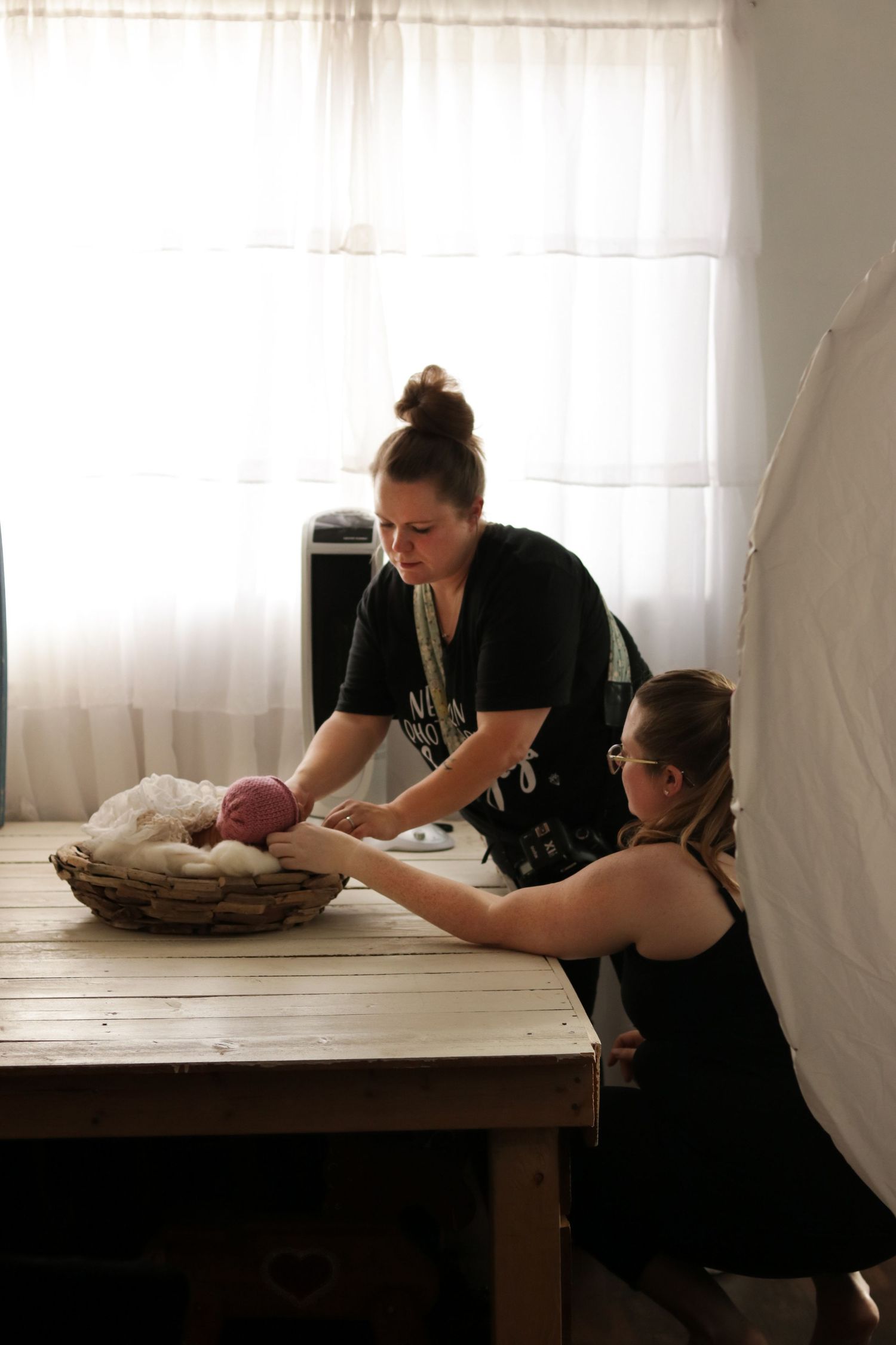 Two people in black clothes work together styling a newborn photo shoot setup on a wooden table near sheer curtains.