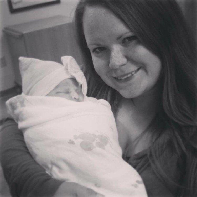 A smiling woman holds a swaddled newborn baby in a black and white hospital photo.