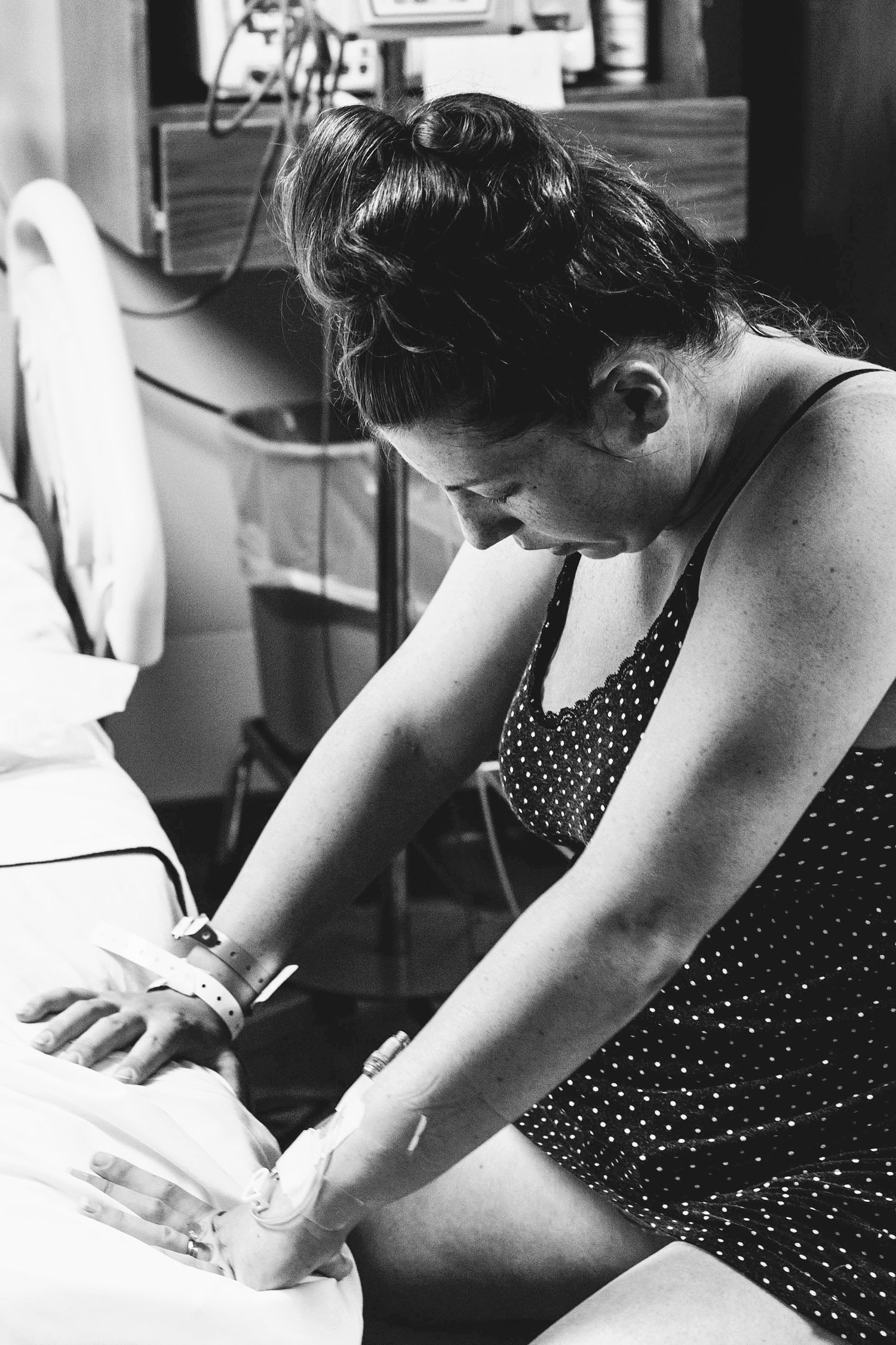 Woman in polka dot dress holds newborn baby's hand in hospital room, black and white birth photography.
