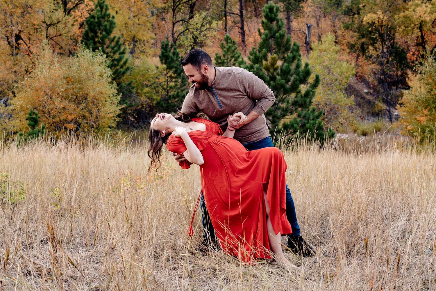 Couple dances romantically in golden autumn field, man dipping woman in red maternity gown.