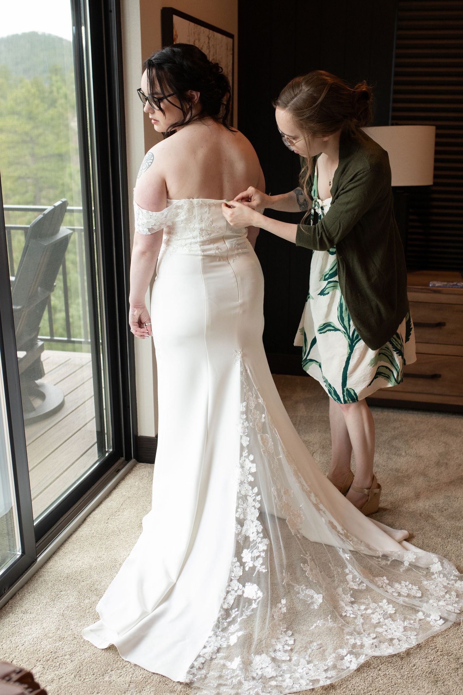 Bridesmaid helping bride with off-shoulder lace train wedding dress near window with mountain view.
