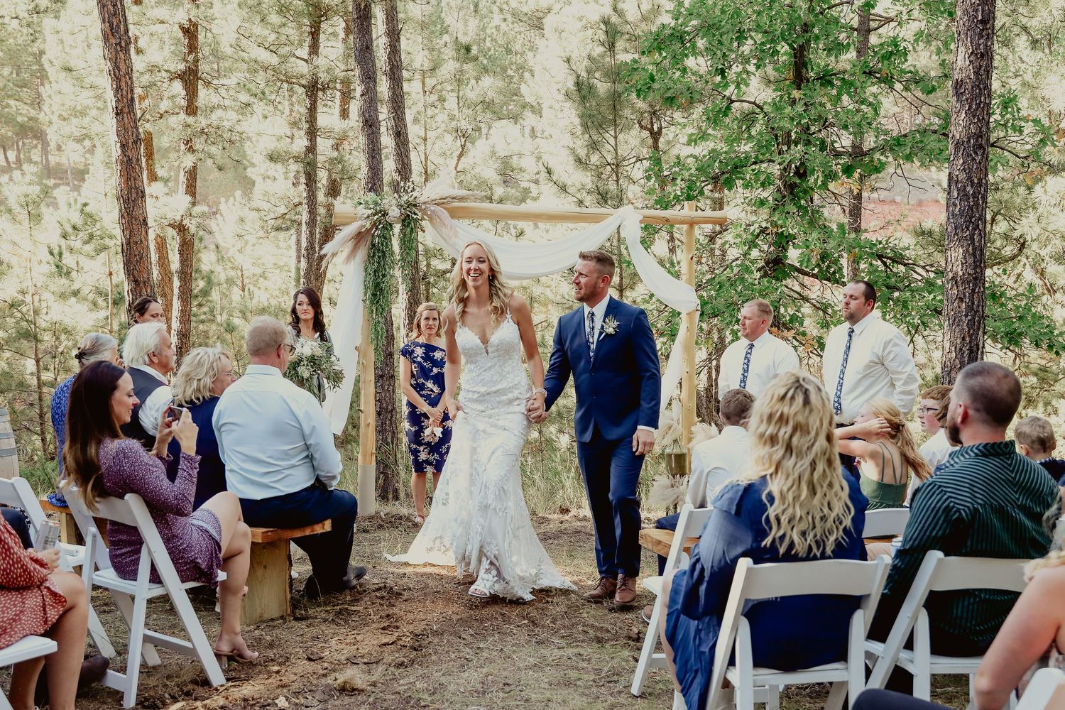Bride and groom walk down outdoor aisle surrounded by guests and tall pine trees at a rustic forest wedding ceremony.