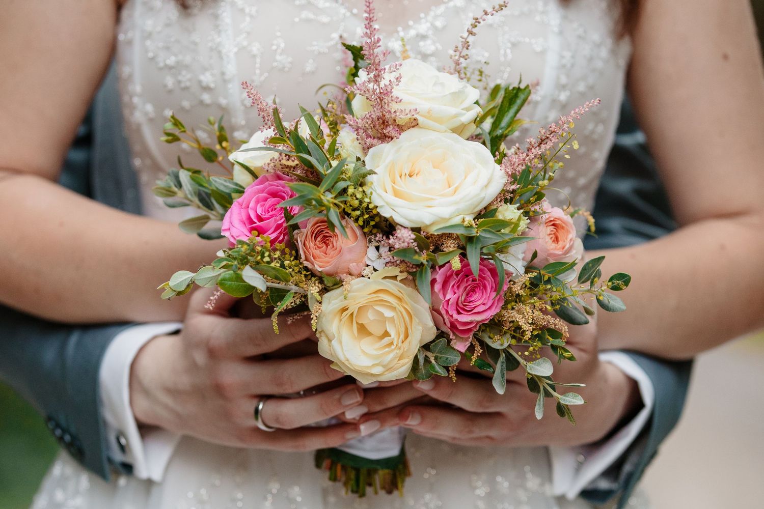 Bride and groom holding a romantic wedding bouquet with pink and cream roses, greenery, and delicate flowers.