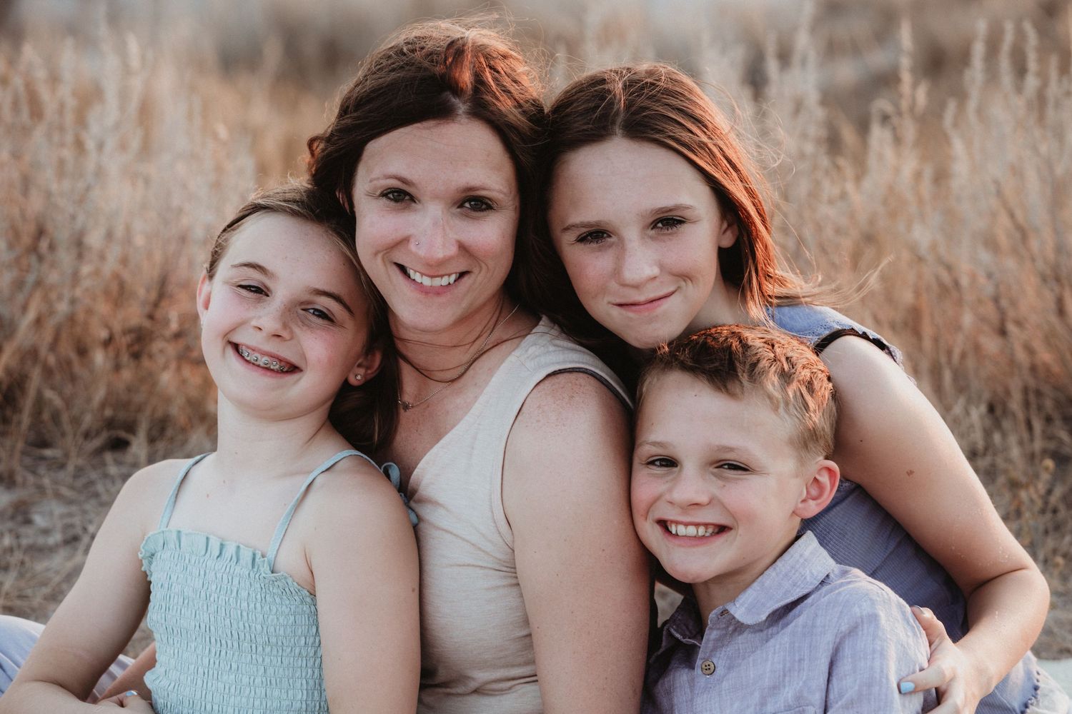 A smiling mother poses outdoors with her three children in a warm golden field during a family portrait session.