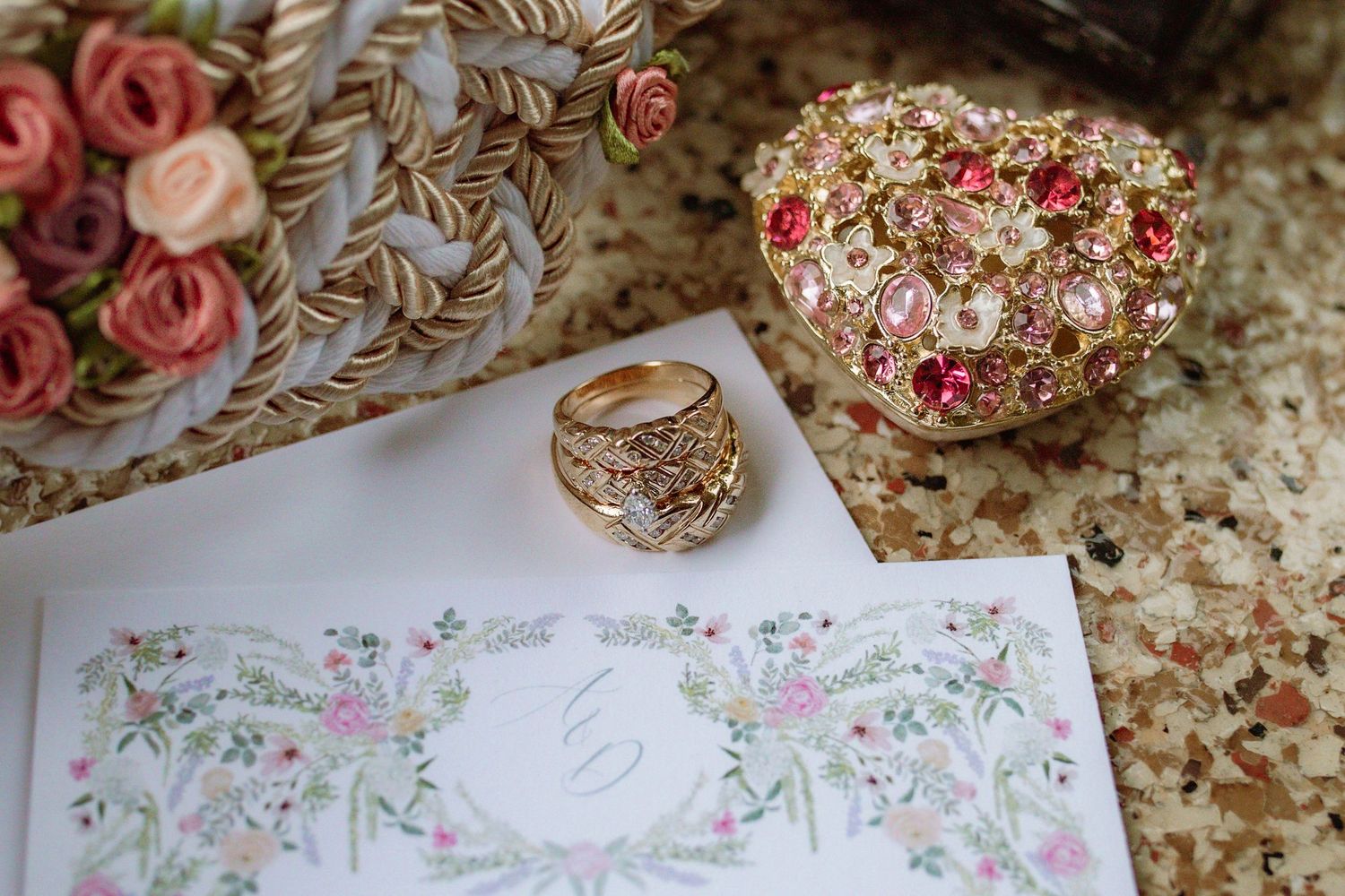 Gold ring with pink and red gemstones beside a floral jewelry box and elegant wedding stationery on a marble surface.