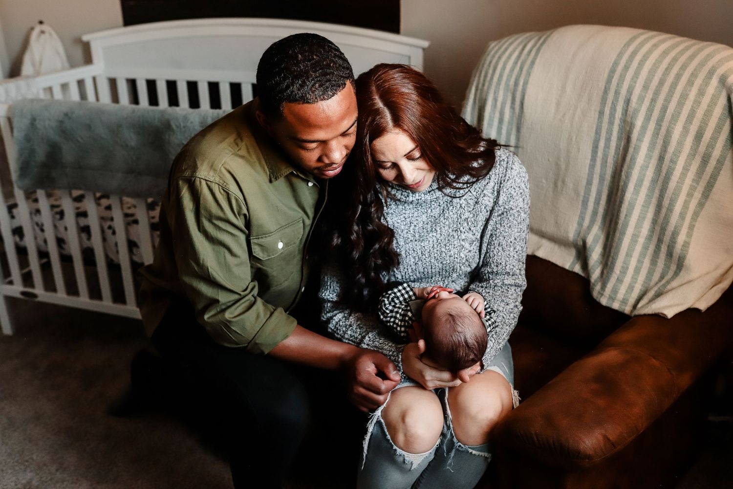 Couple sits together on nursery floor admiring their newborn baby in front of a white crib.