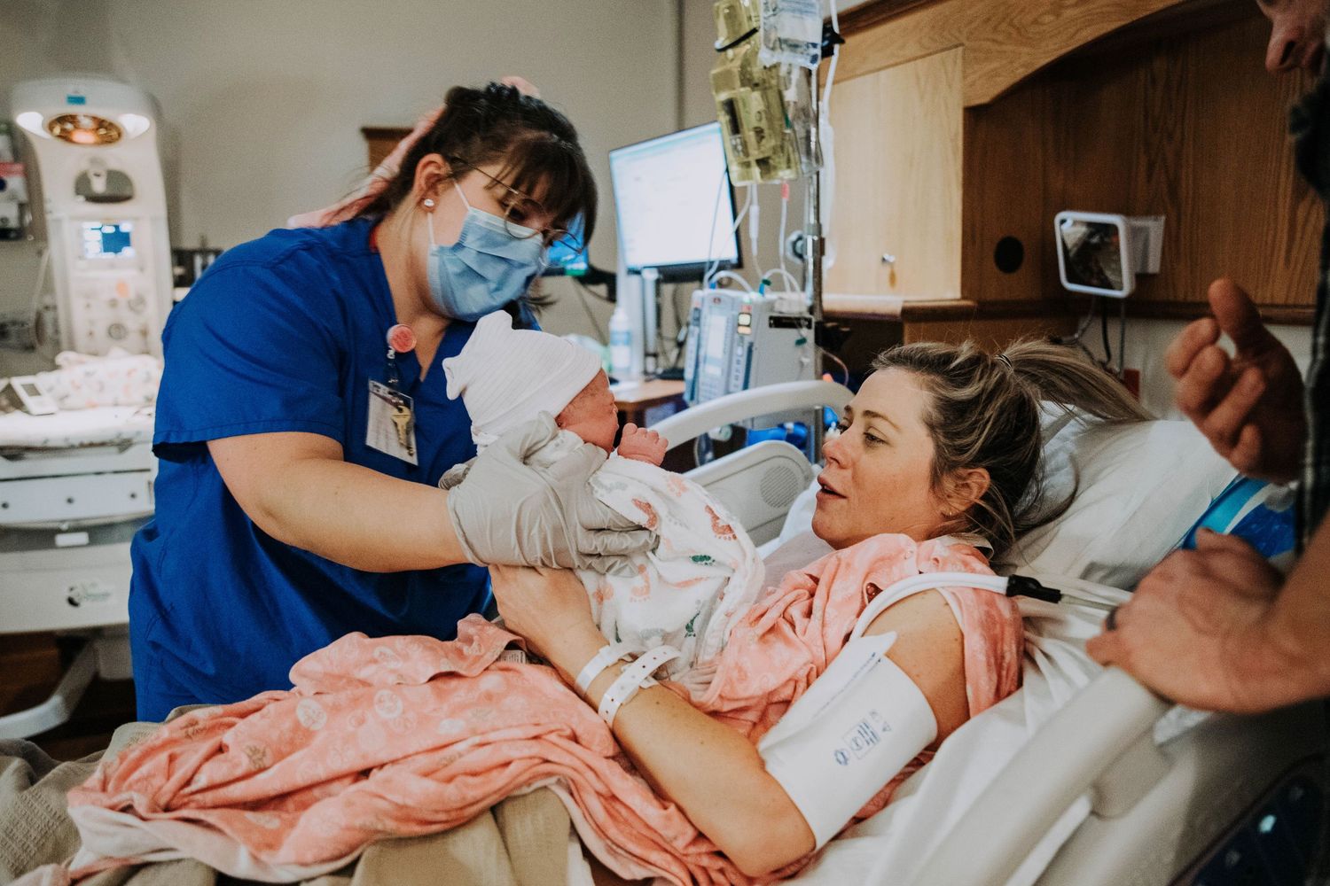 Nurse in blue scrubs handing newborn baby to smiling mother in hospital delivery room.