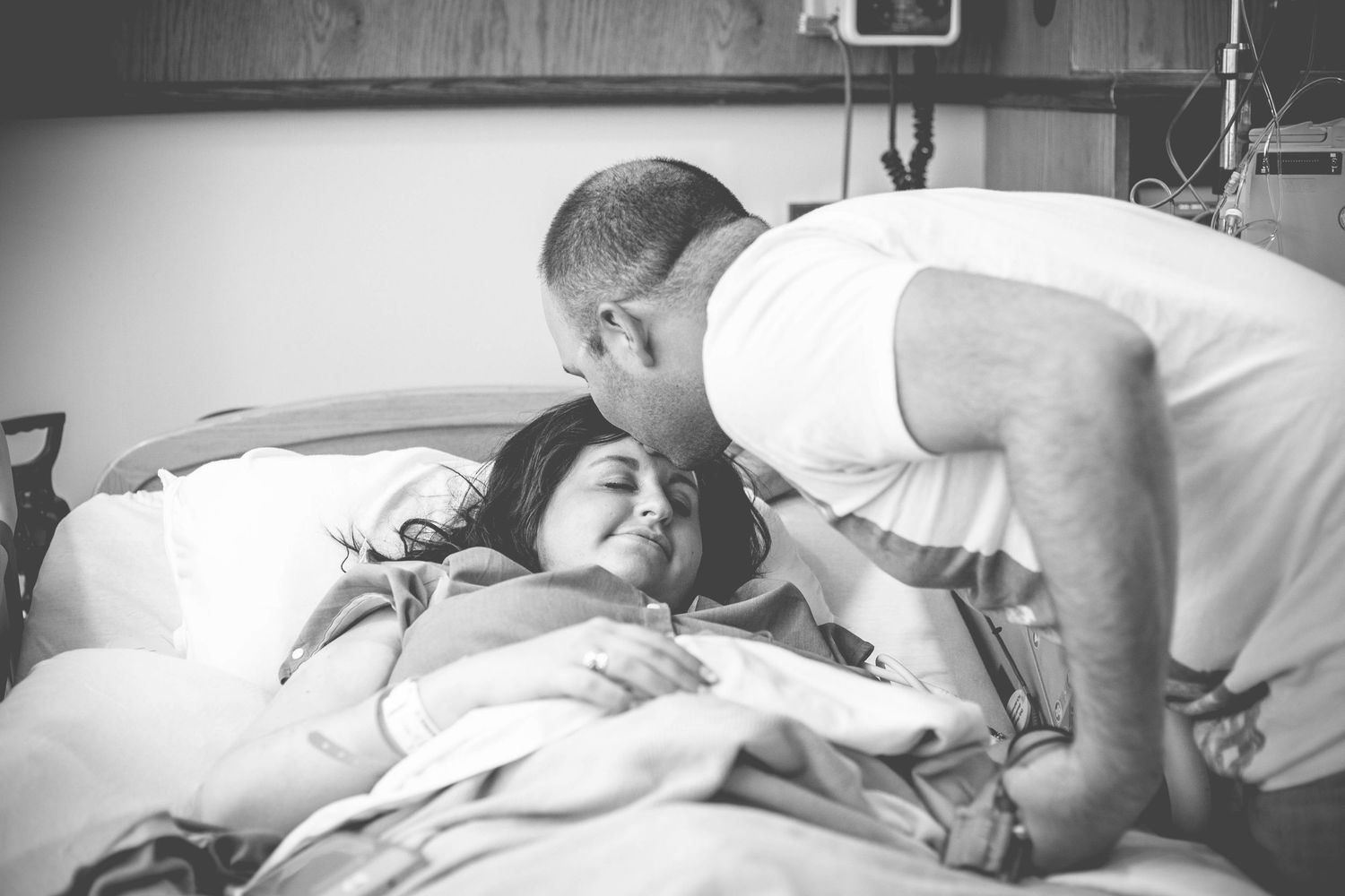 A tender black and white hospital moment as a man kisses his partner lying in a hospital bed.