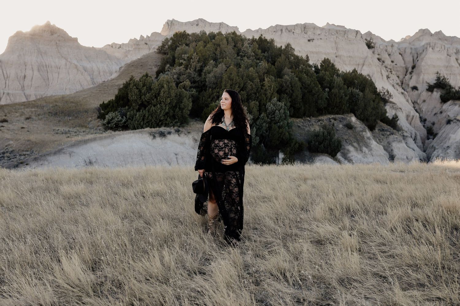 Pregnant woman in black floral dress walking through field with badlands background.