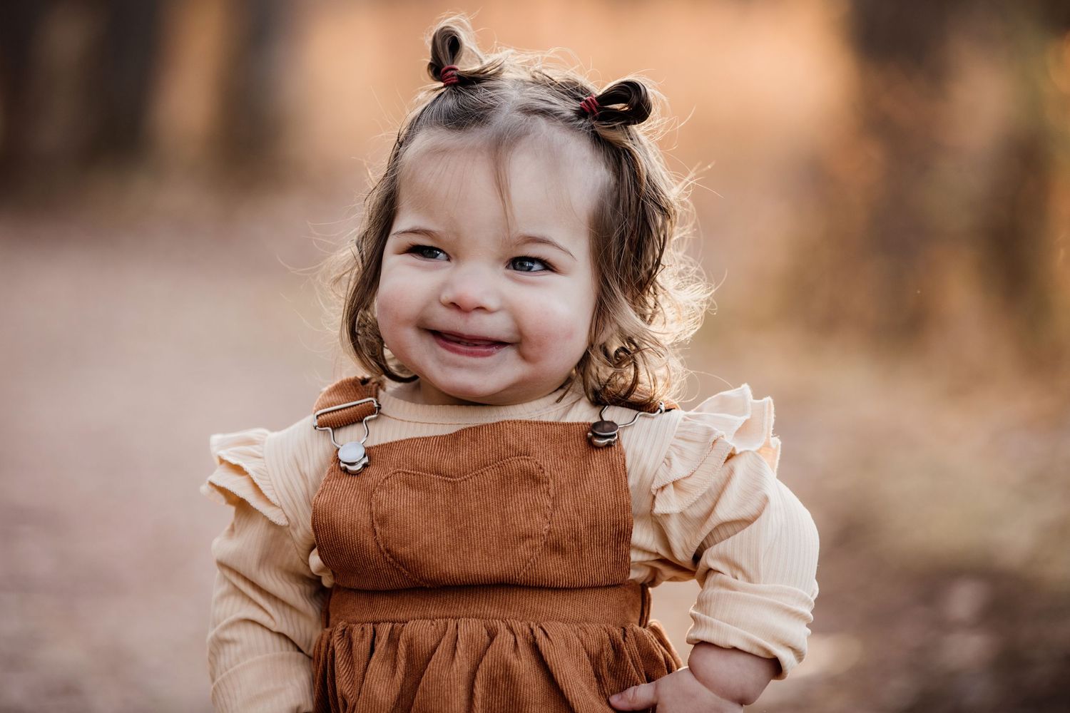 Smiling toddler girl in brown overalls and cream top with bear ear hair clips outdoors in warm autumn setting.