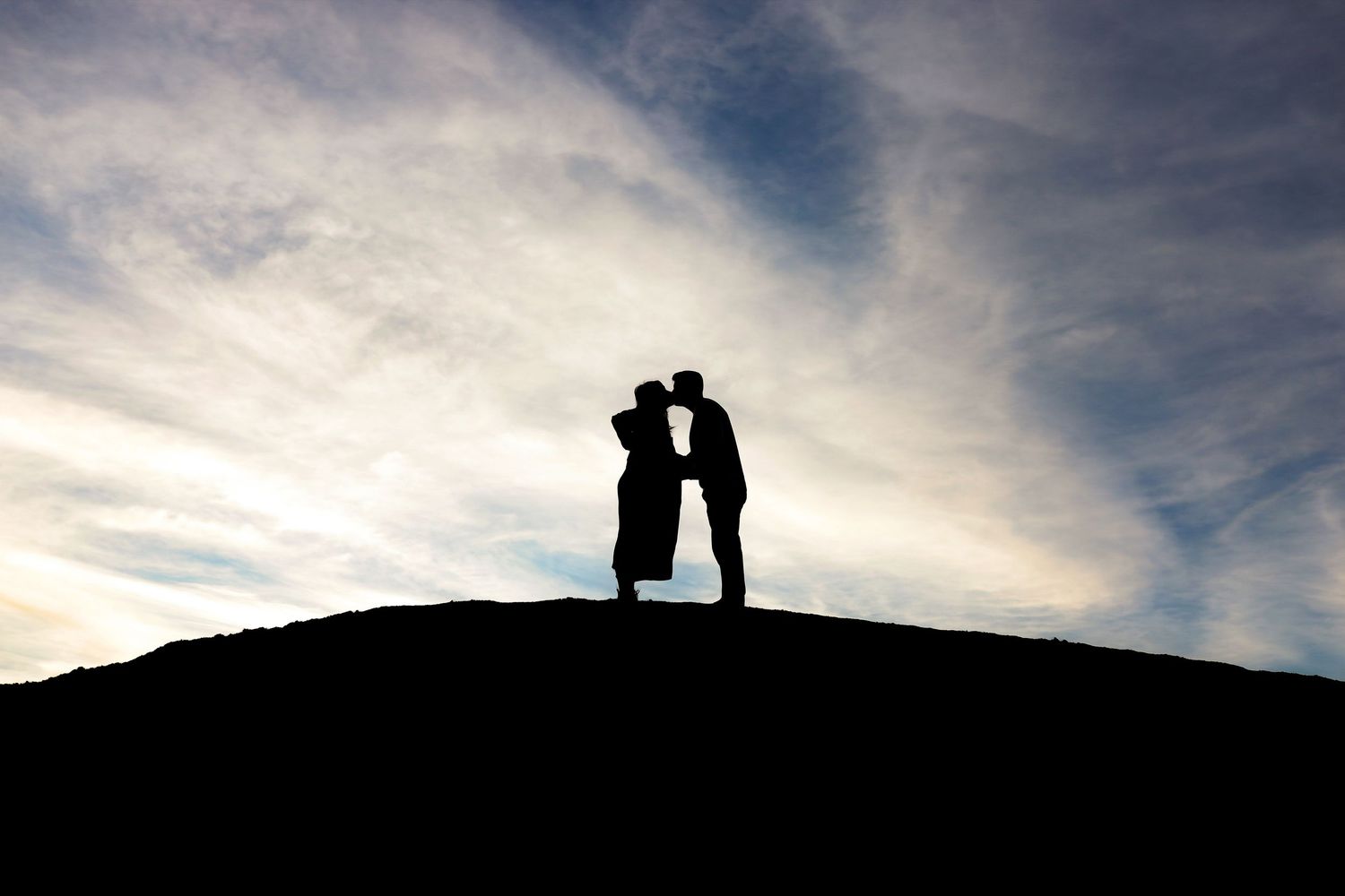 Romantic silhouette of couple kissing on hilltop against dramatic cloudy sky.