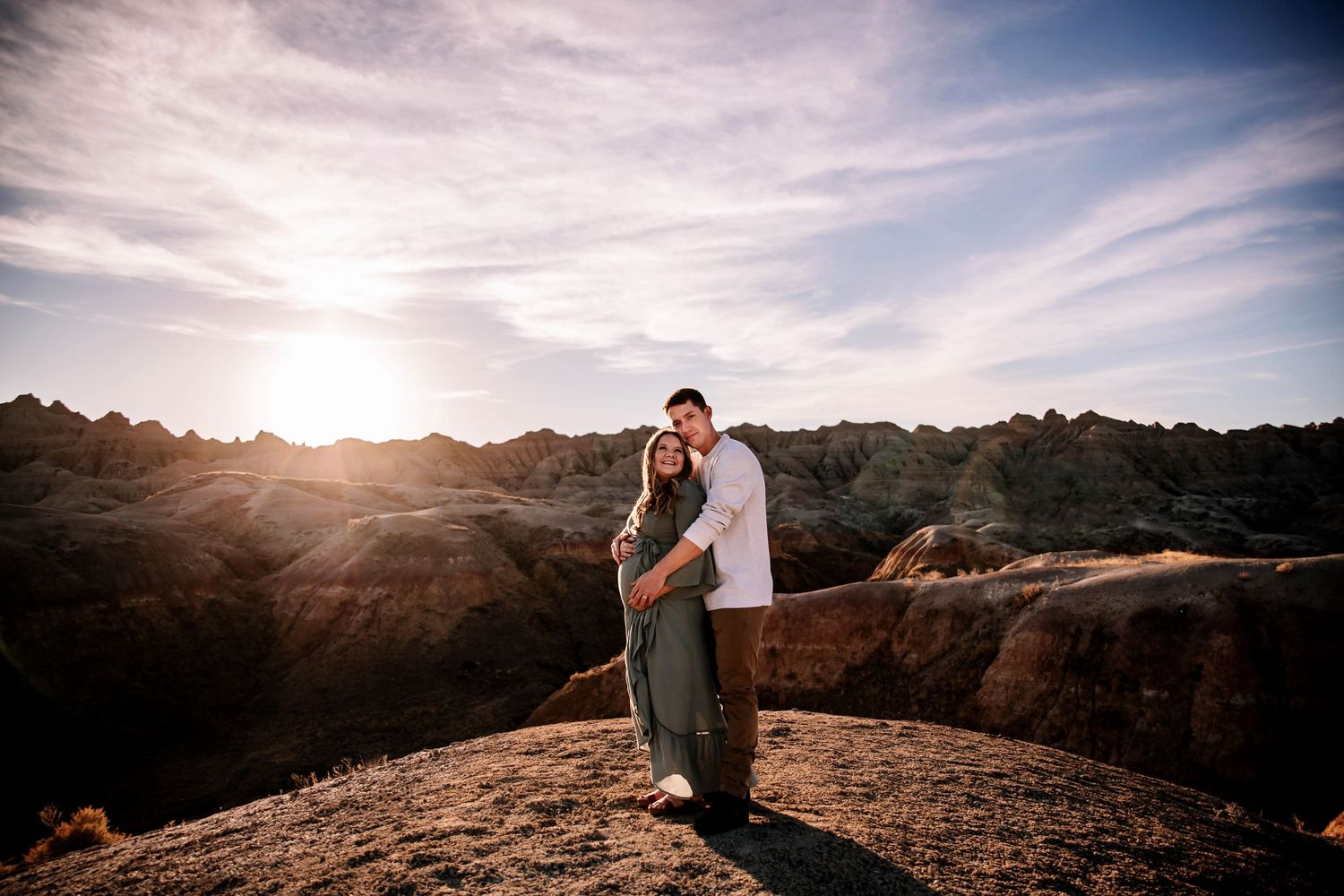 Couple embraces at golden hour atop rocky badlands overlook under dramatic cloudy sky.
