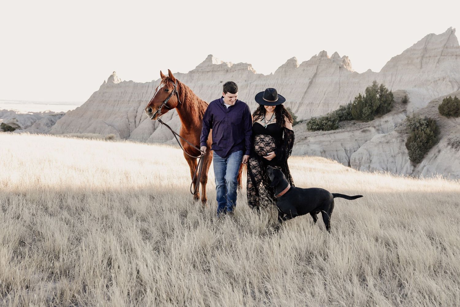 Couple walking with chestnut horse and black Labrador through tall grass in badlands landscape.