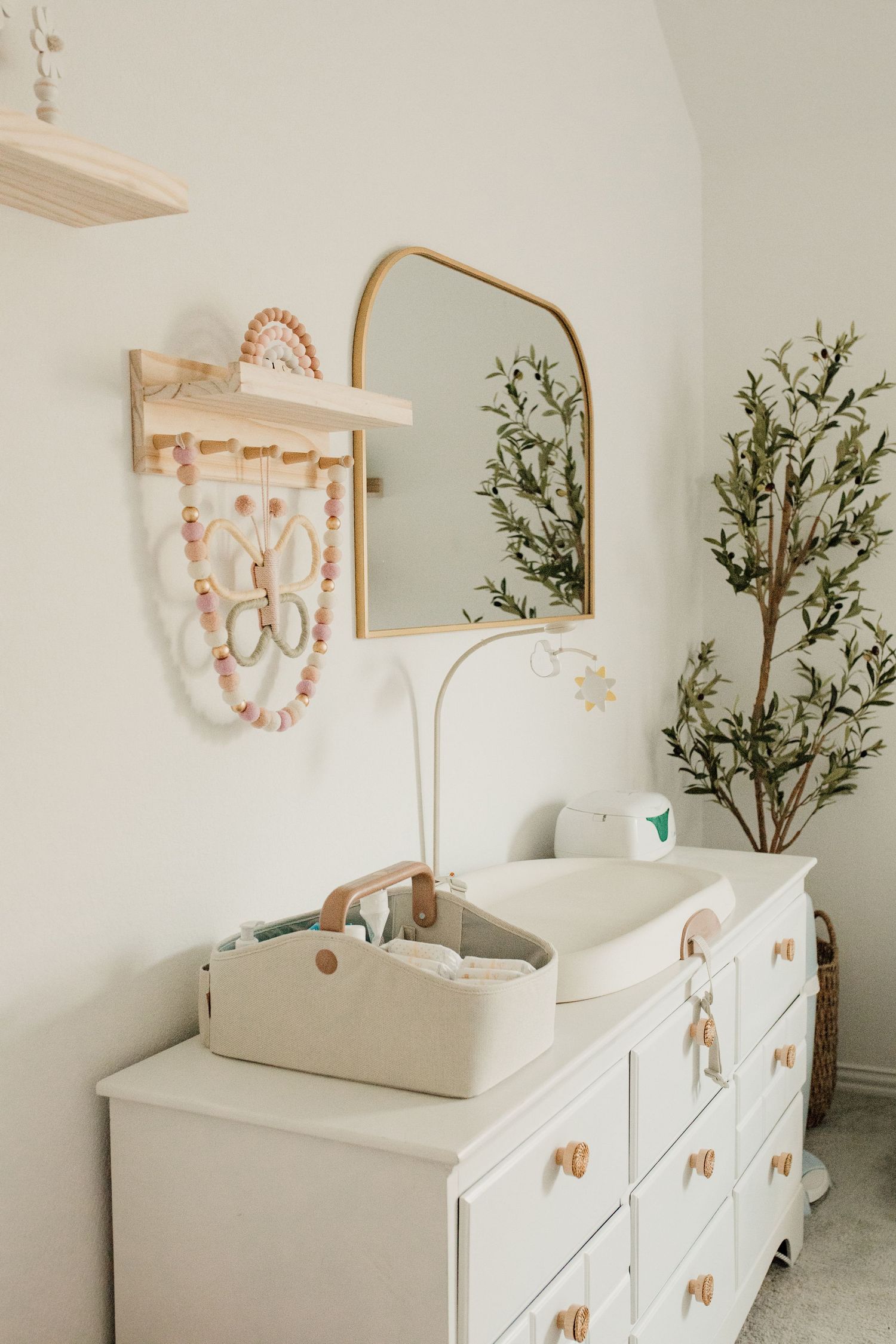 White nursery dresser with changing pad, gold arch mirror, wooden toy shelf, and decorative jewelry display on wall.
