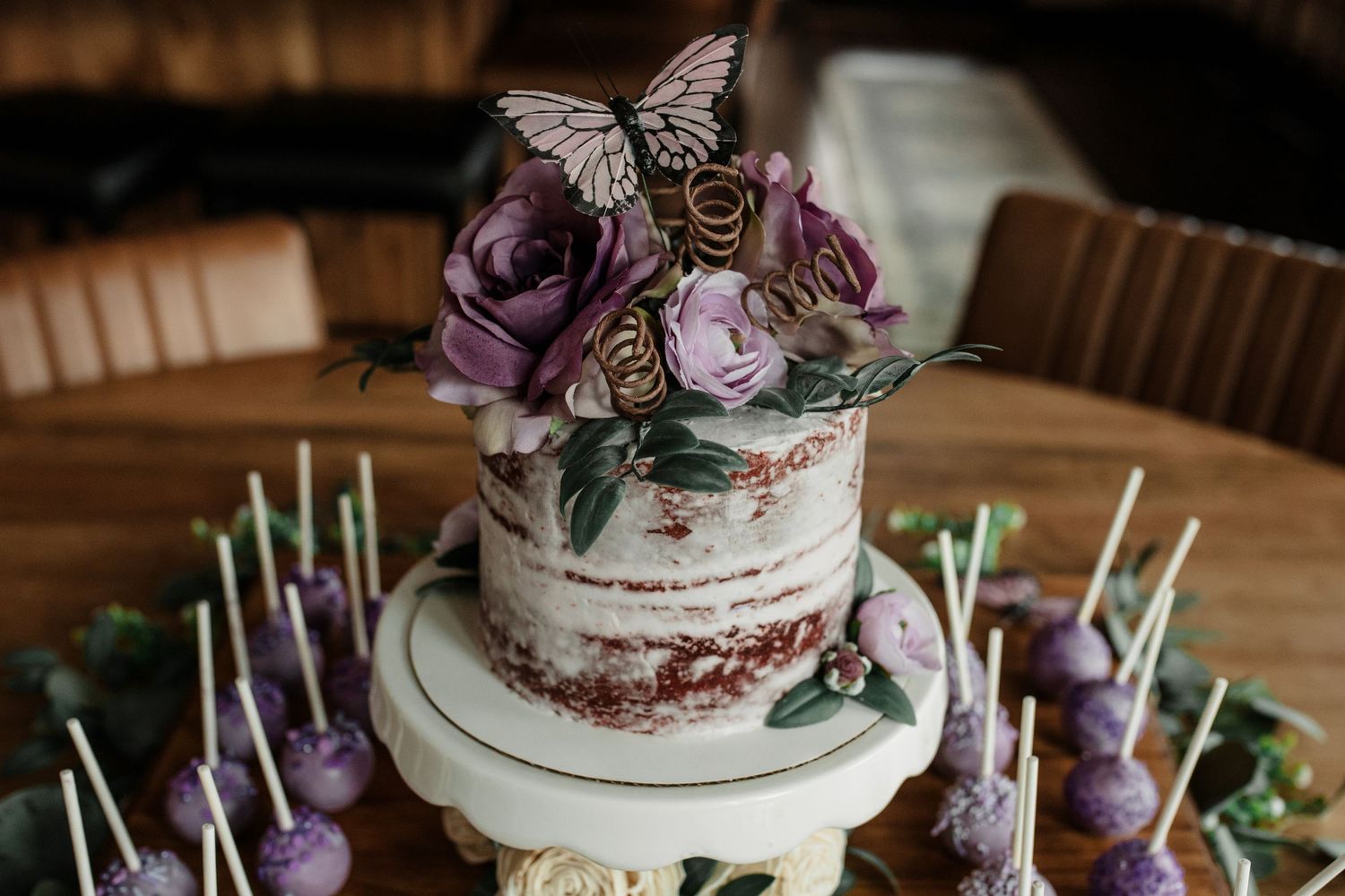 Semi-naked wedding cake with purple roses and butterfly topper, surrounded by purple cake pops on sticks.