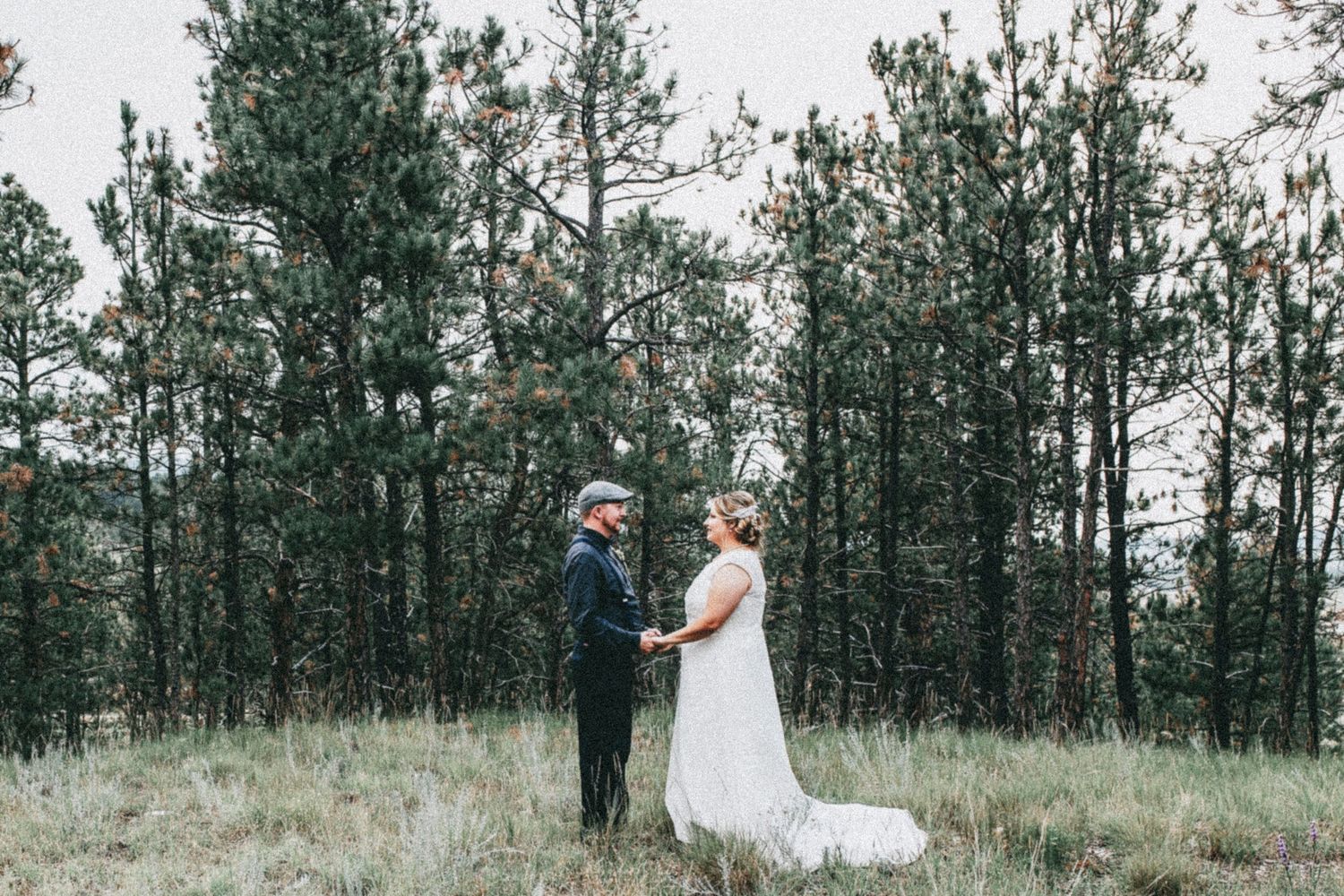 Couple holds hands during outdoor wedding ceremony in a forest meadow, bride in white gown with veil, groom in dark suit.