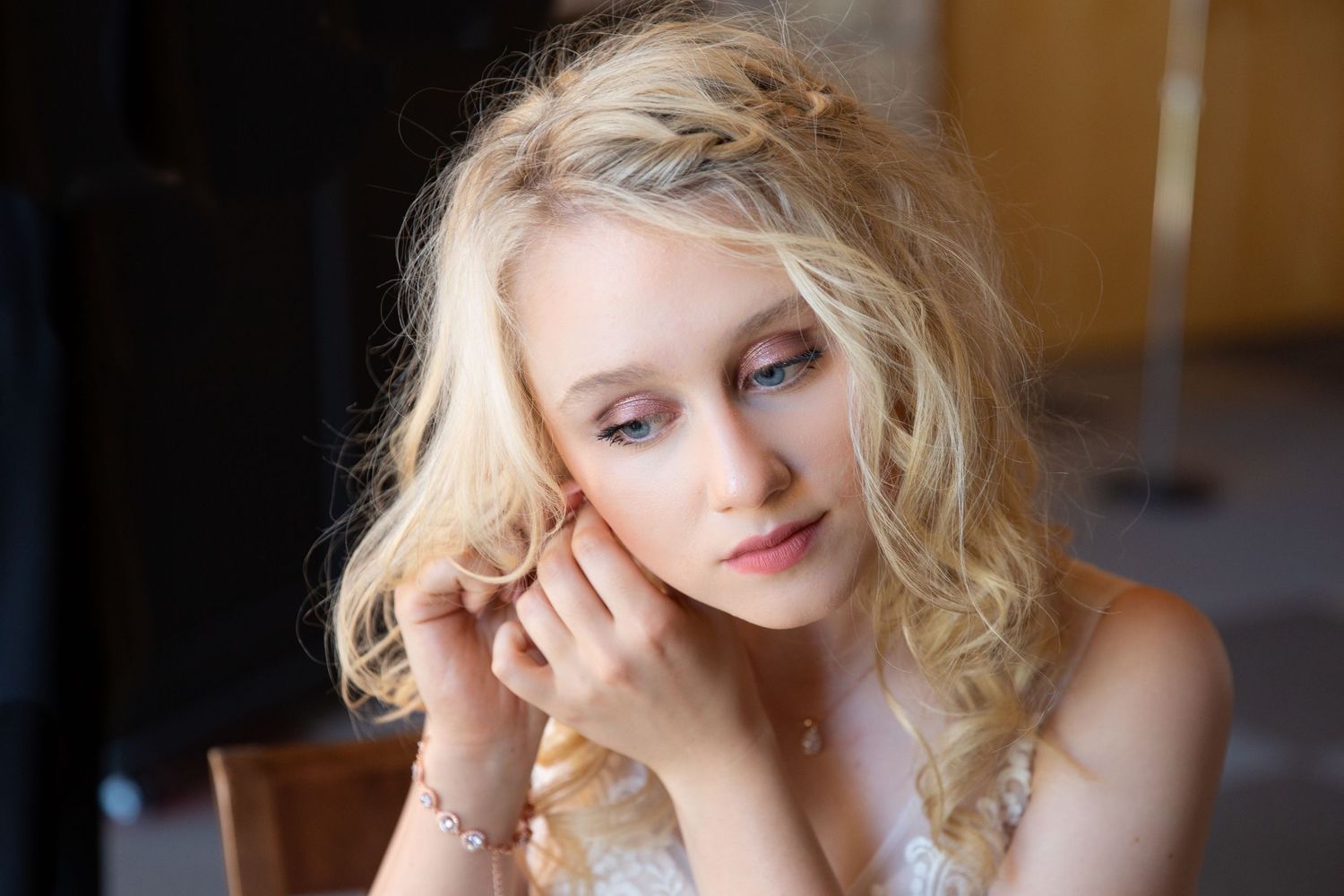 Young blonde woman with curly hair and braided updo, wearing delicate jewelry, gazing down thoughtfully.