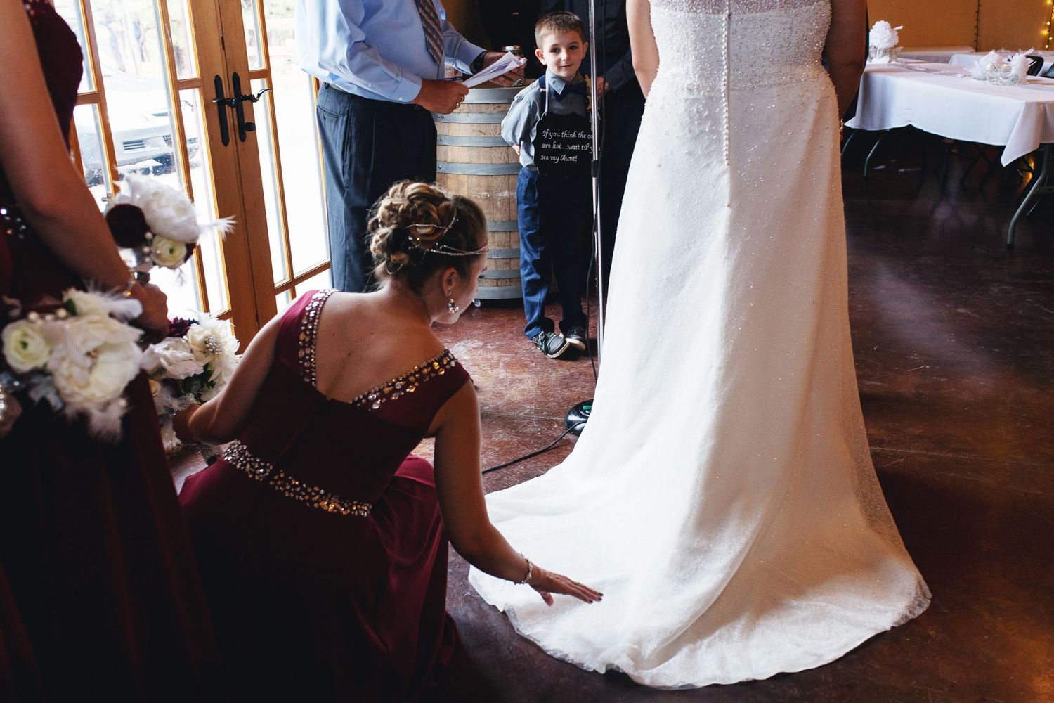 Bridesmaid in burgundy gown adjusting bride's white wedding dress train while a young boy watches indoors.