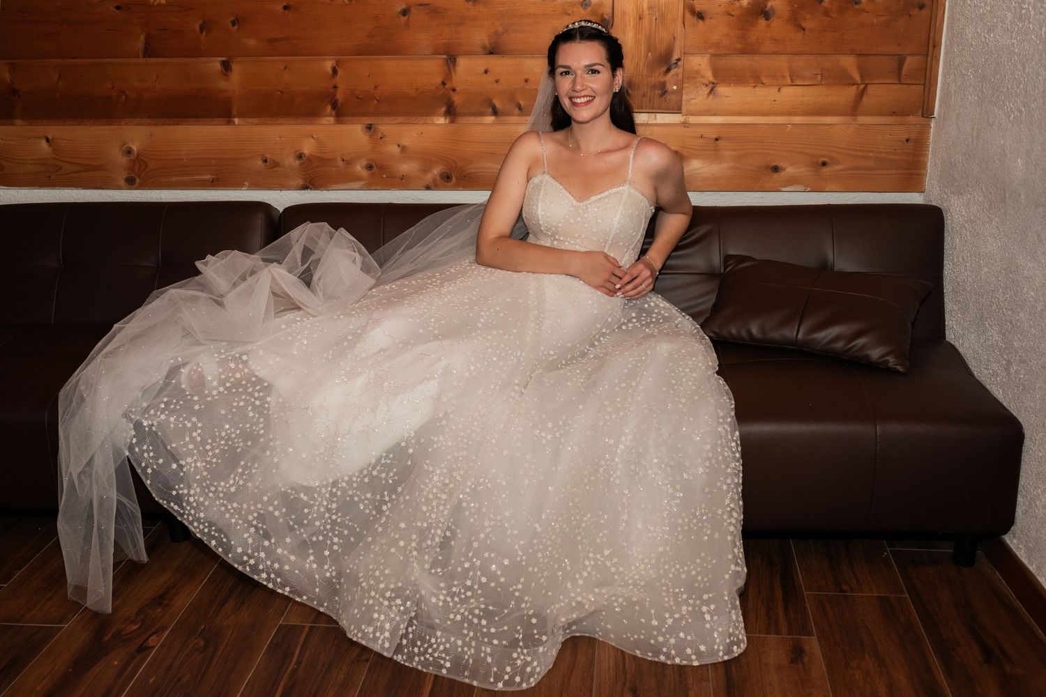 Bride in a sparkling ball gown with tiara sits gracefully on a brown leather sofa in a wood-paneled room.