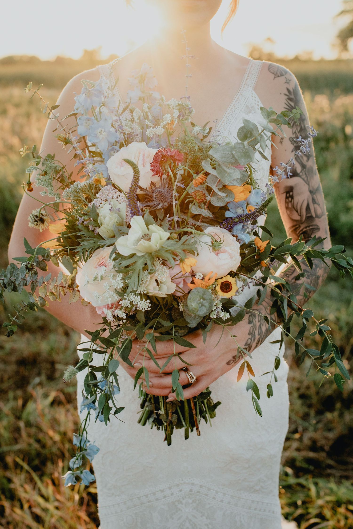 Tattooed bride holding a lush wildflower wedding bouquet with white, pink, and blue blooms in golden sunset light.