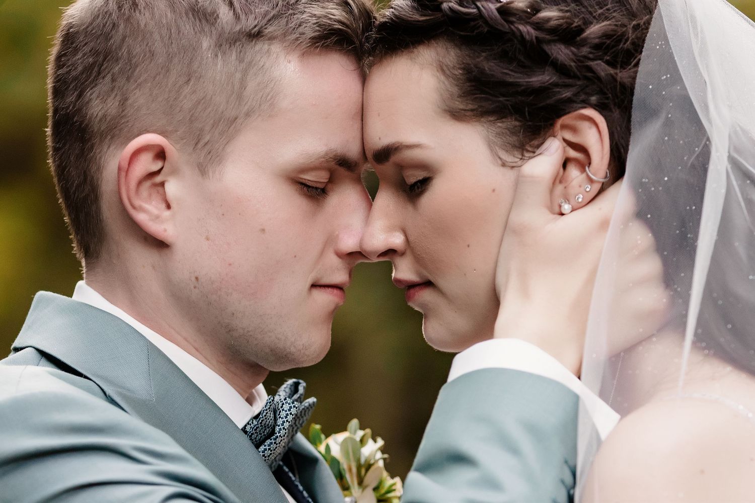 Bride and groom share an intimate forehead touch moment under a flowing veil outdoors.