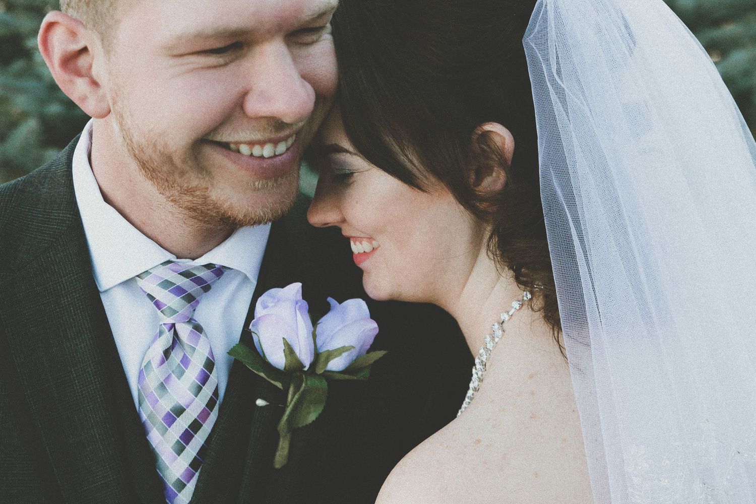 A smiling groom in a suit with purple boutonniere leans close to his bride in a white veil on their wedding day.