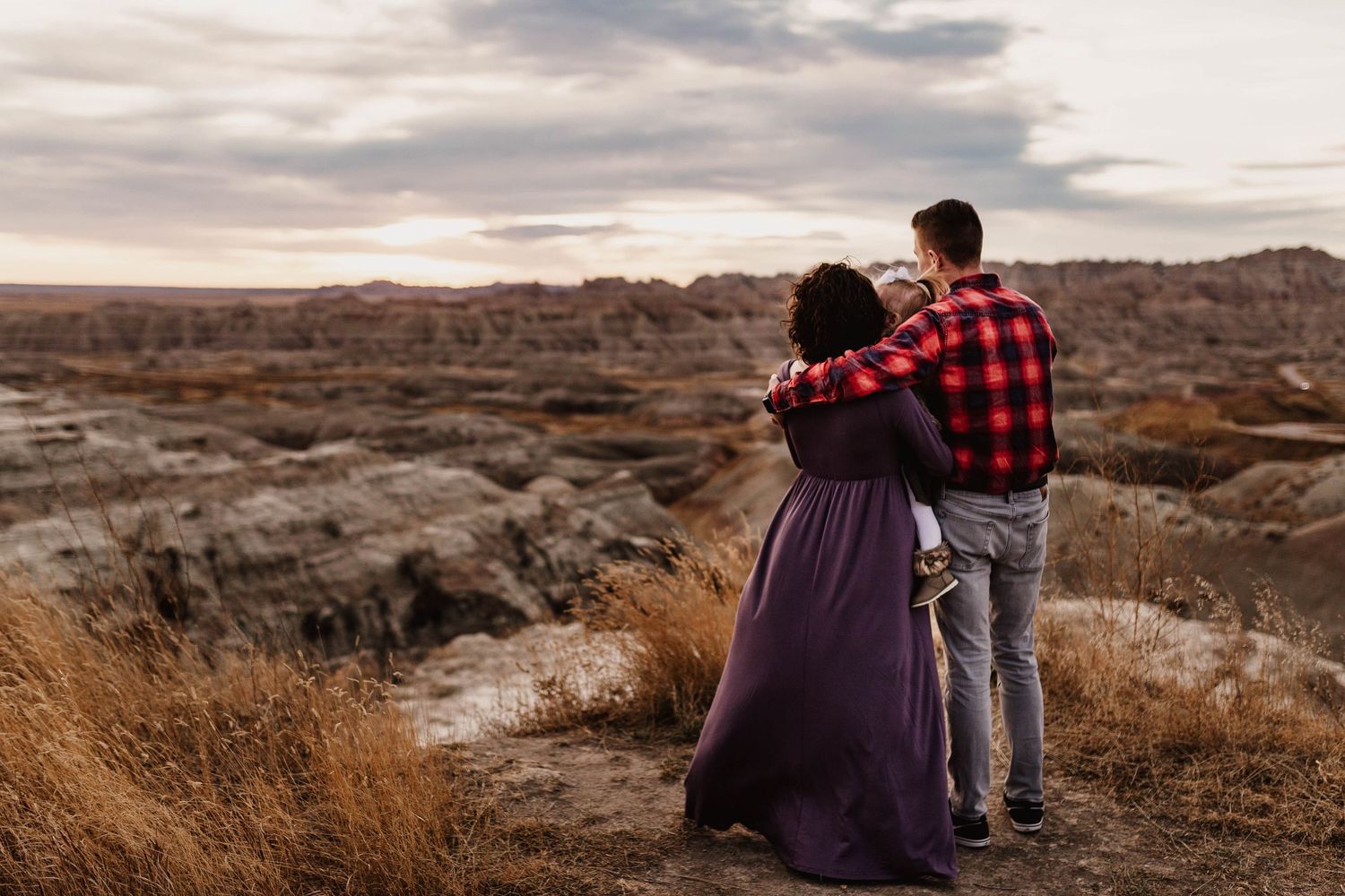 Couple embracing while overlooking dramatic badlands landscape at sunset in purple dress and plaid shirt.